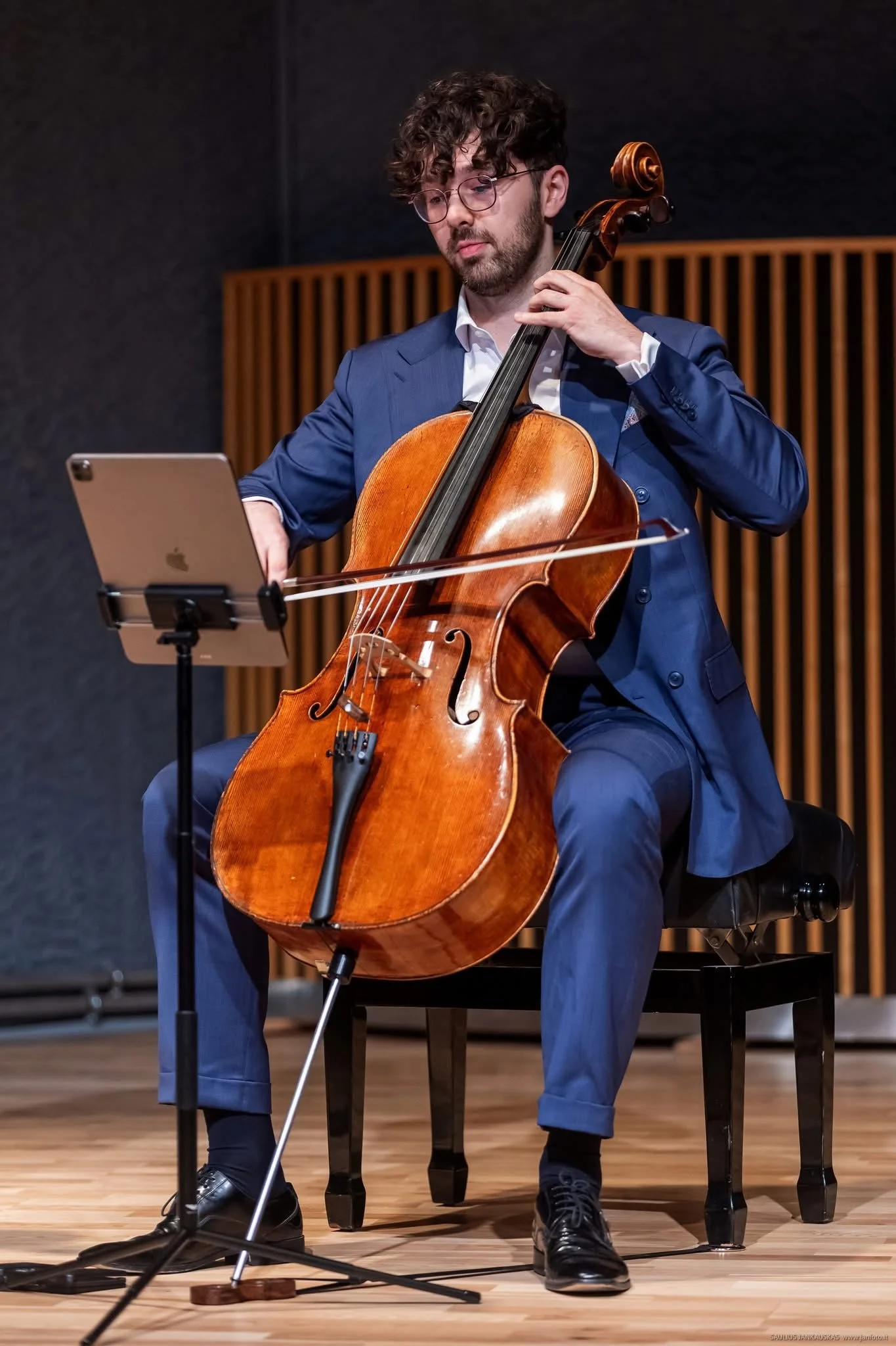 A man in a blue suit playing a cello on stage with a music stand holding a tablet nearby.