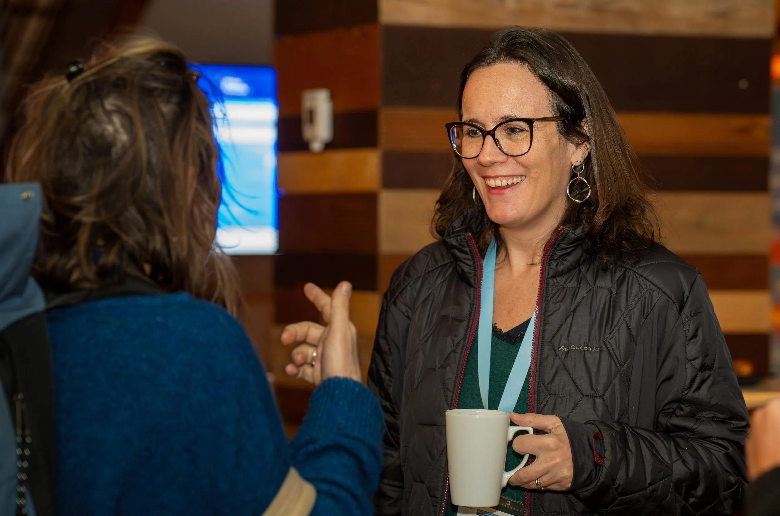 Two women engaging in conversation, one holding a white mug, in a warm indoor setting with wooden paneling. EU Delegation Seminar on Human Rights 2025, Democracy & Gender Equality Brussels