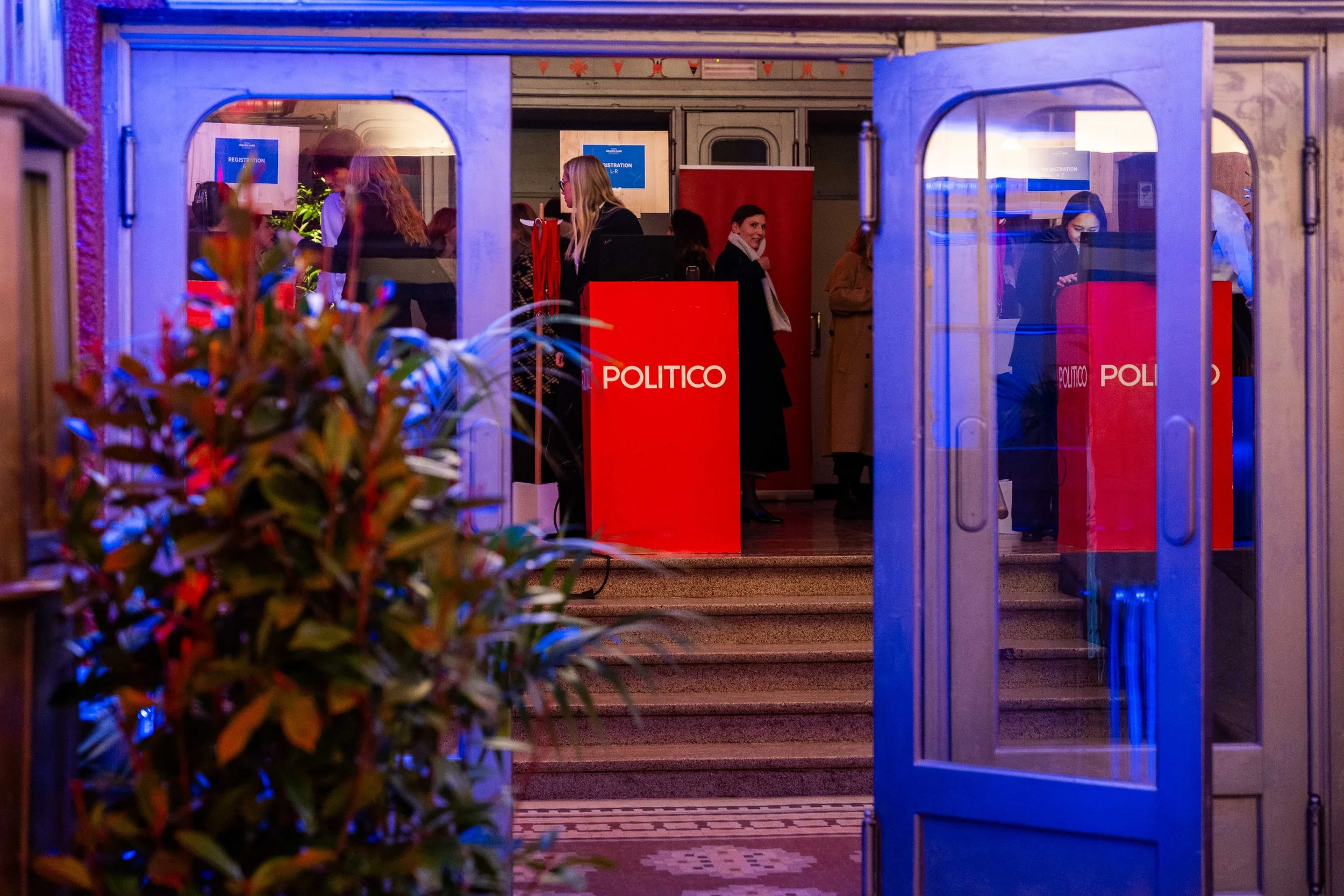 People entering an event at a venue with red 'POLITICO' logos on the stands, viewed through an open glass door with a potted plant in the foreground. Summit Photographer Brussels.