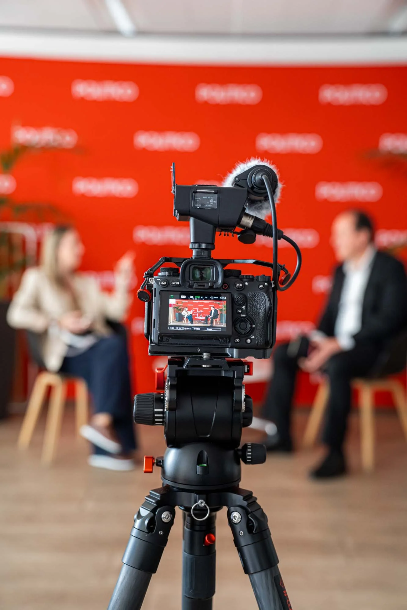 A professional camera on a tripod positioned in front of two people seated on chairs, blurred in the background with a red backdrop.
