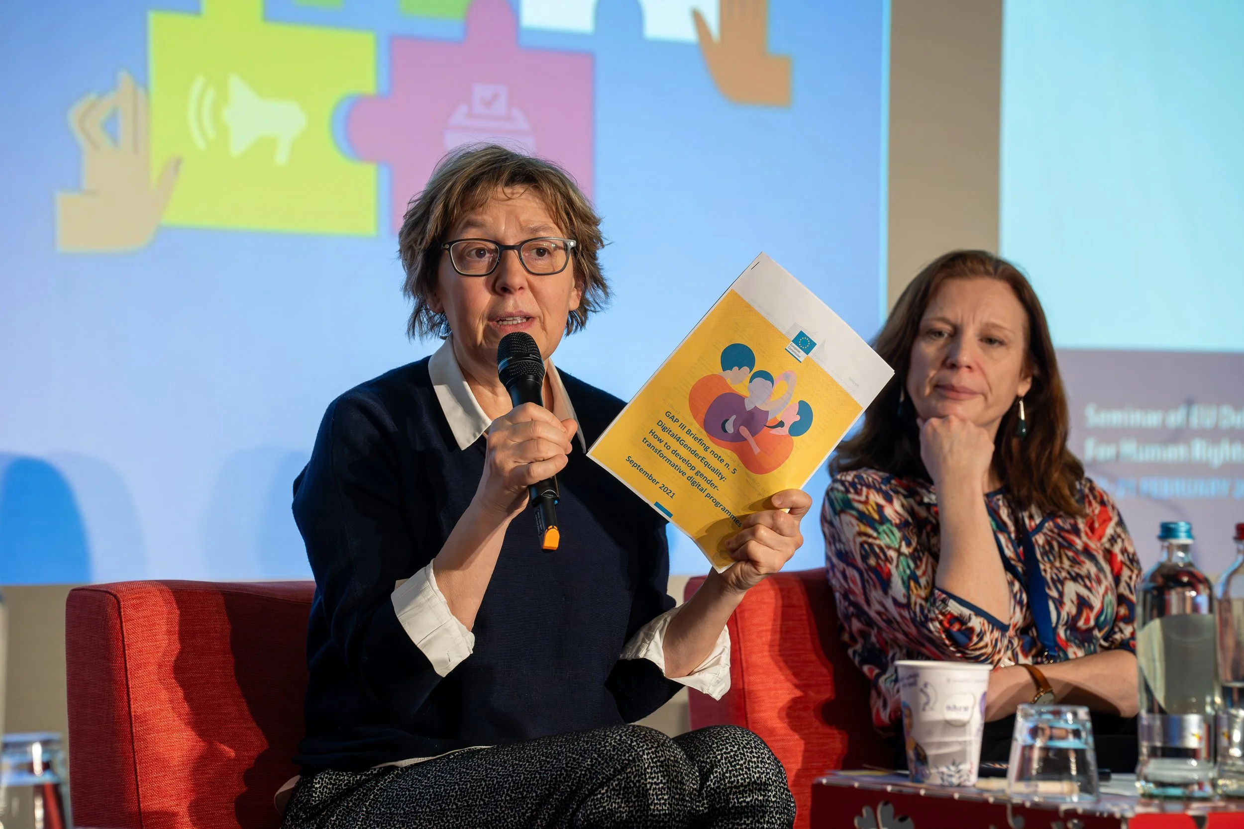 A woman with glasses and short hair speaking into a microphone during a panel discussion. She is holding a brochure or booklet. Another woman with long hair and patterned clothing sits next to her with her hand near her face. There are water bottles 