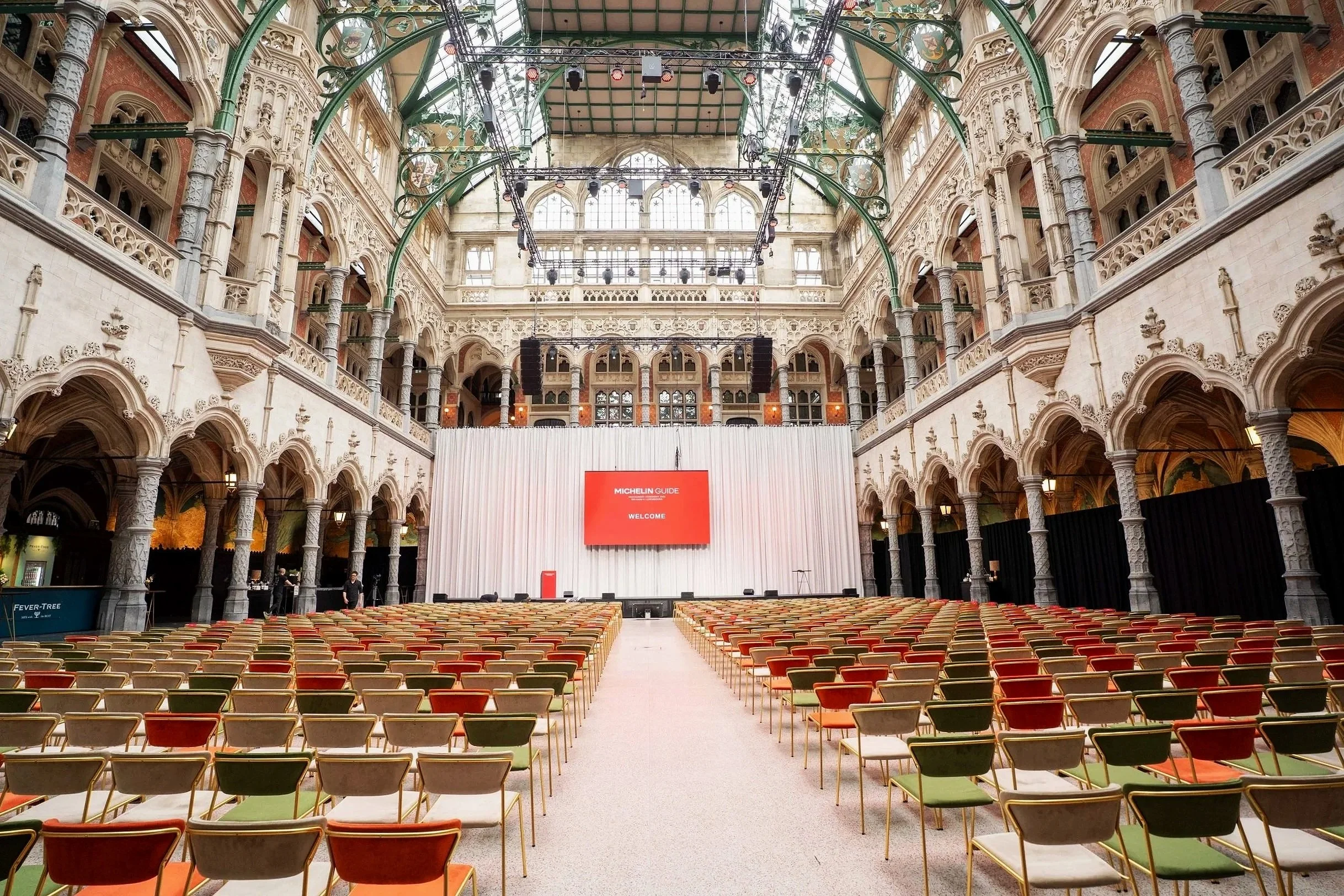 A grand, ornate hall with multiple levels of balconies and detailed architectural decorations. A central stage with a large red screen reading 'Michelin Guide Welcome' is set up, surrounded by rows of empty chairs in front.