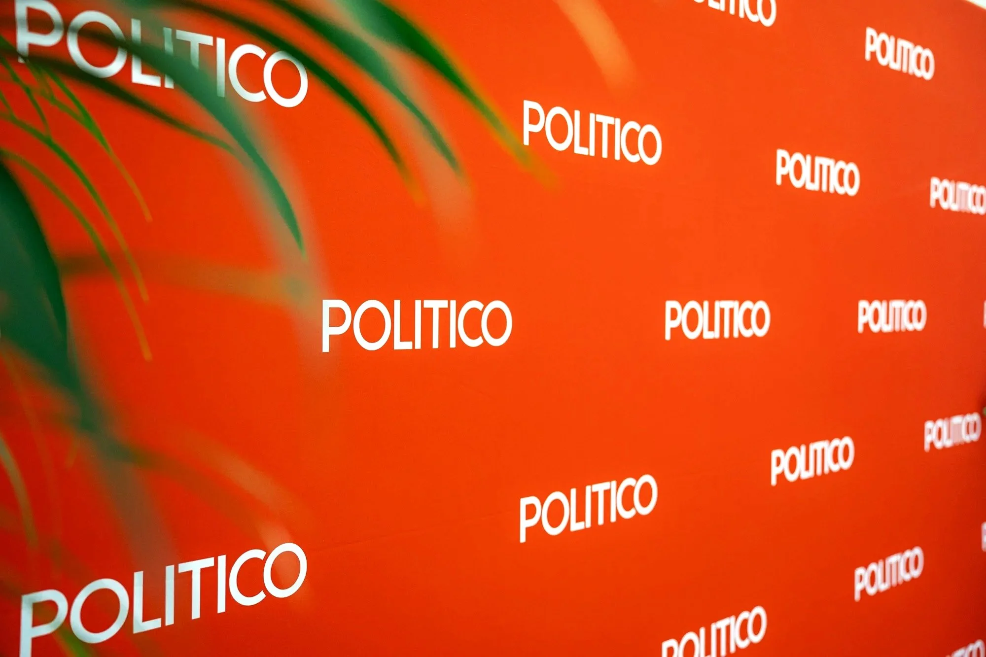 A bright orange background with multiple white words 'POLITICO' repeatedly printed across it, and some green shadows or streaks from the left side. Politico Event Photography Brussels.