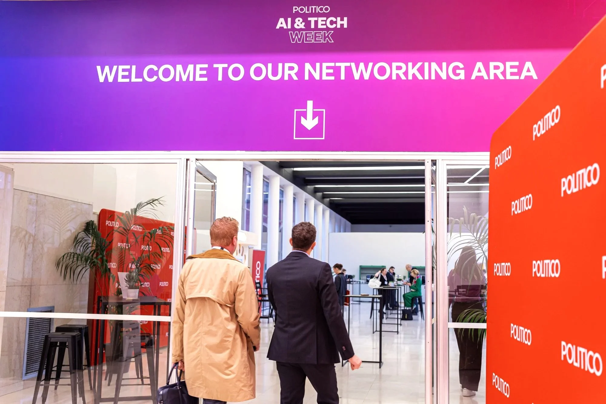 People walking into a networking area at POLITICO AI & Tech Week, with purple and orange banners, tables, and attendees inside. Politico Event Photography Brussels.