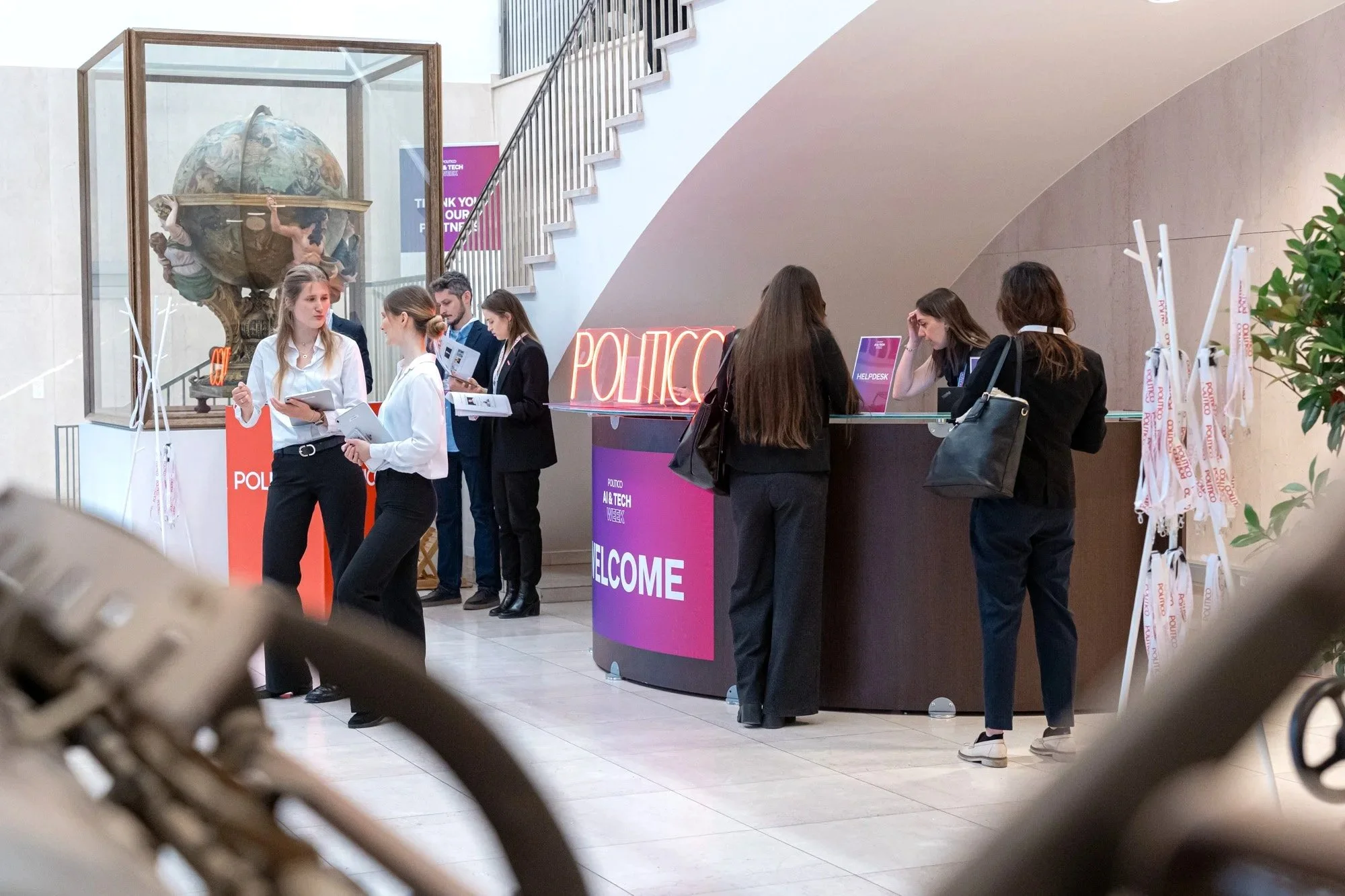 A group of people standing at a reception desk with a neon 'POLITICO' sign and a large globe sculpture behind a glass case in a modern, indoor setting. Politico Event Photography Brussels.