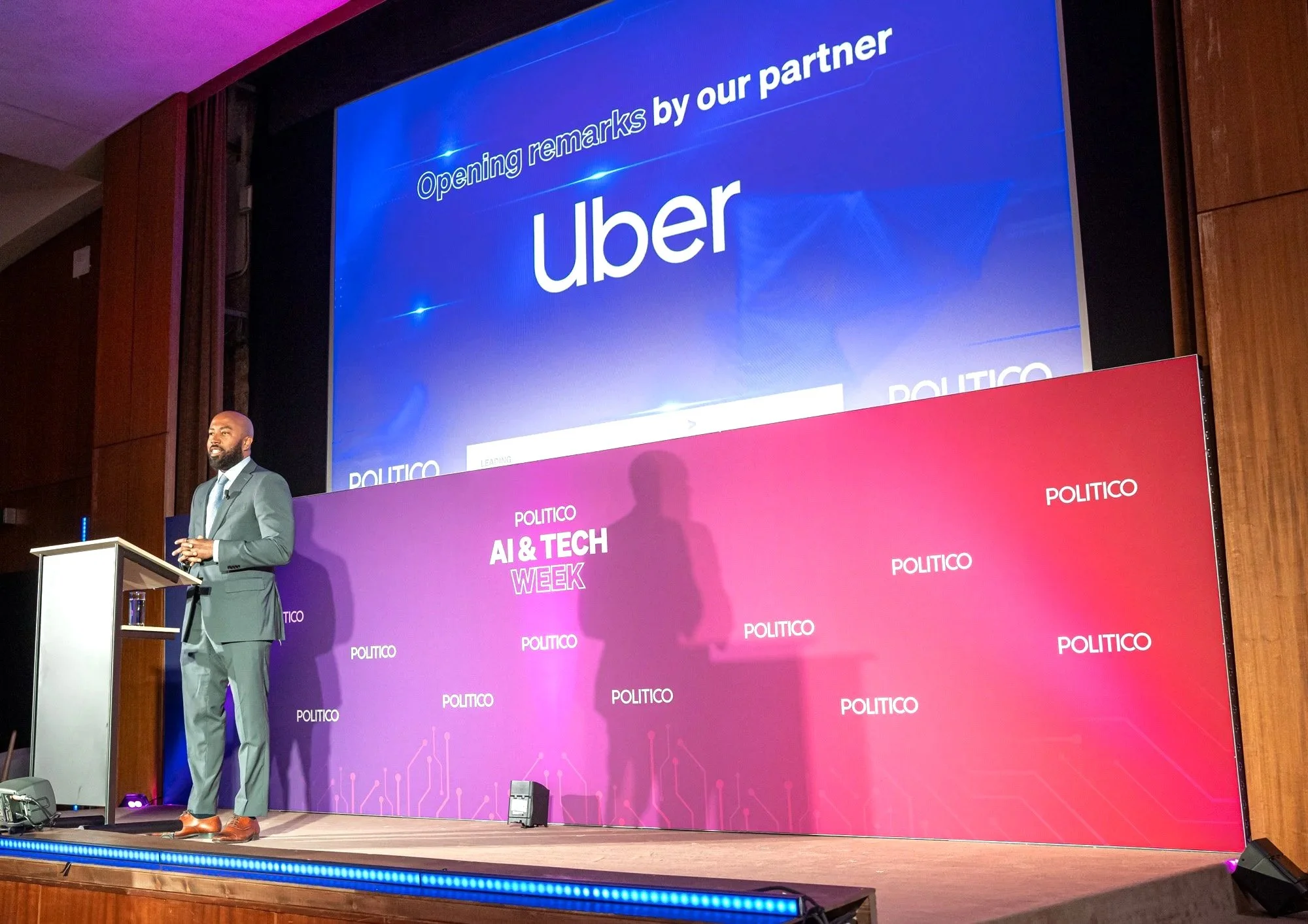 A man in a gray suit speaking at a podium on stage during a tech conference with a large screen behind him displaying UBER branding and the phrase "Opening remarks by our partner." The stage has a purple and pink themed banner with "POLITICO" written