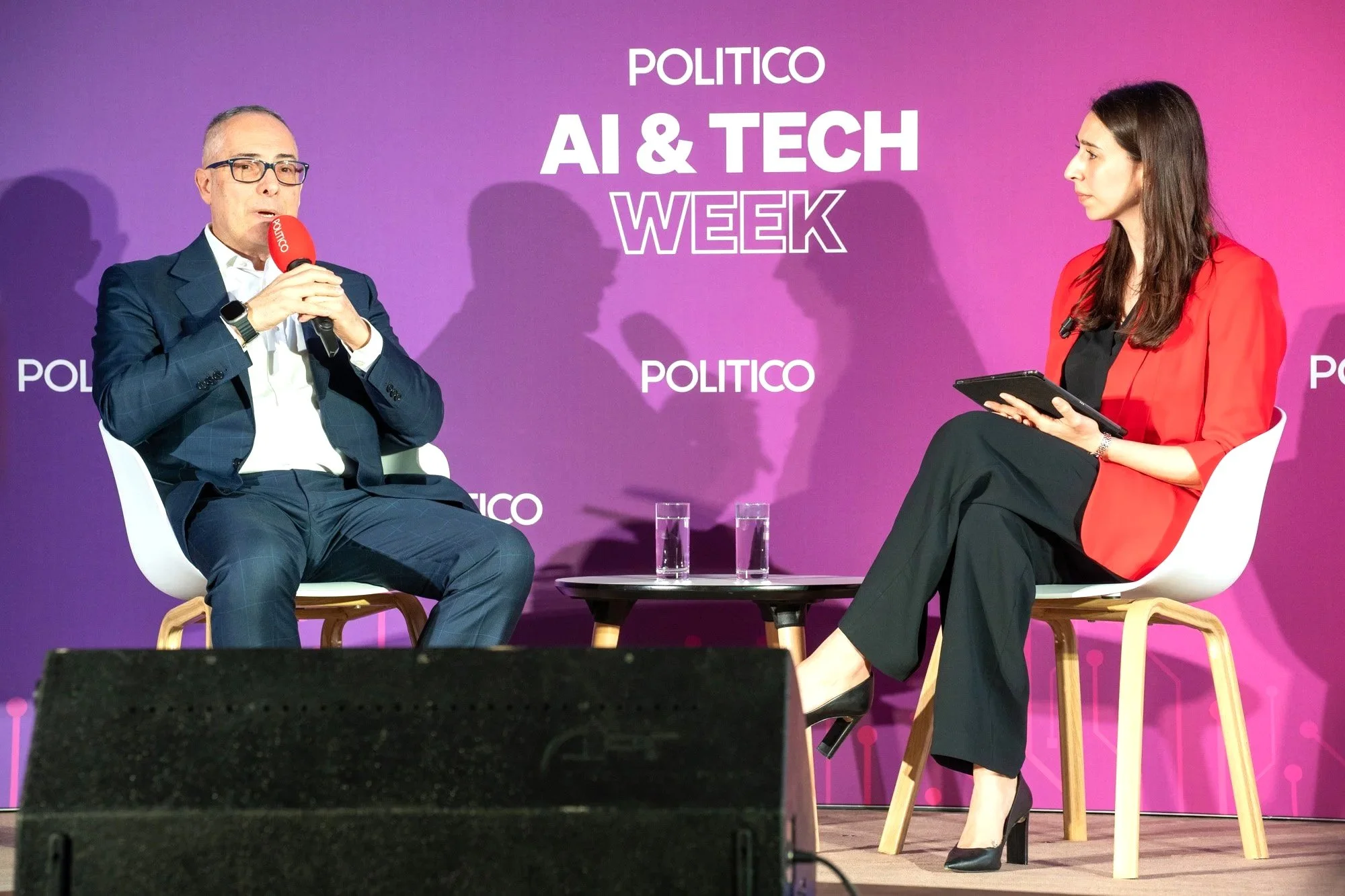 A man and a woman sit on stage during an AI and Tech Week event organized by Politico. The man, on the left, wears glasses, a dark suit, and a white shirt, holding a red microphone. The woman, on the right, wears a red blazer and black pants, and hol
