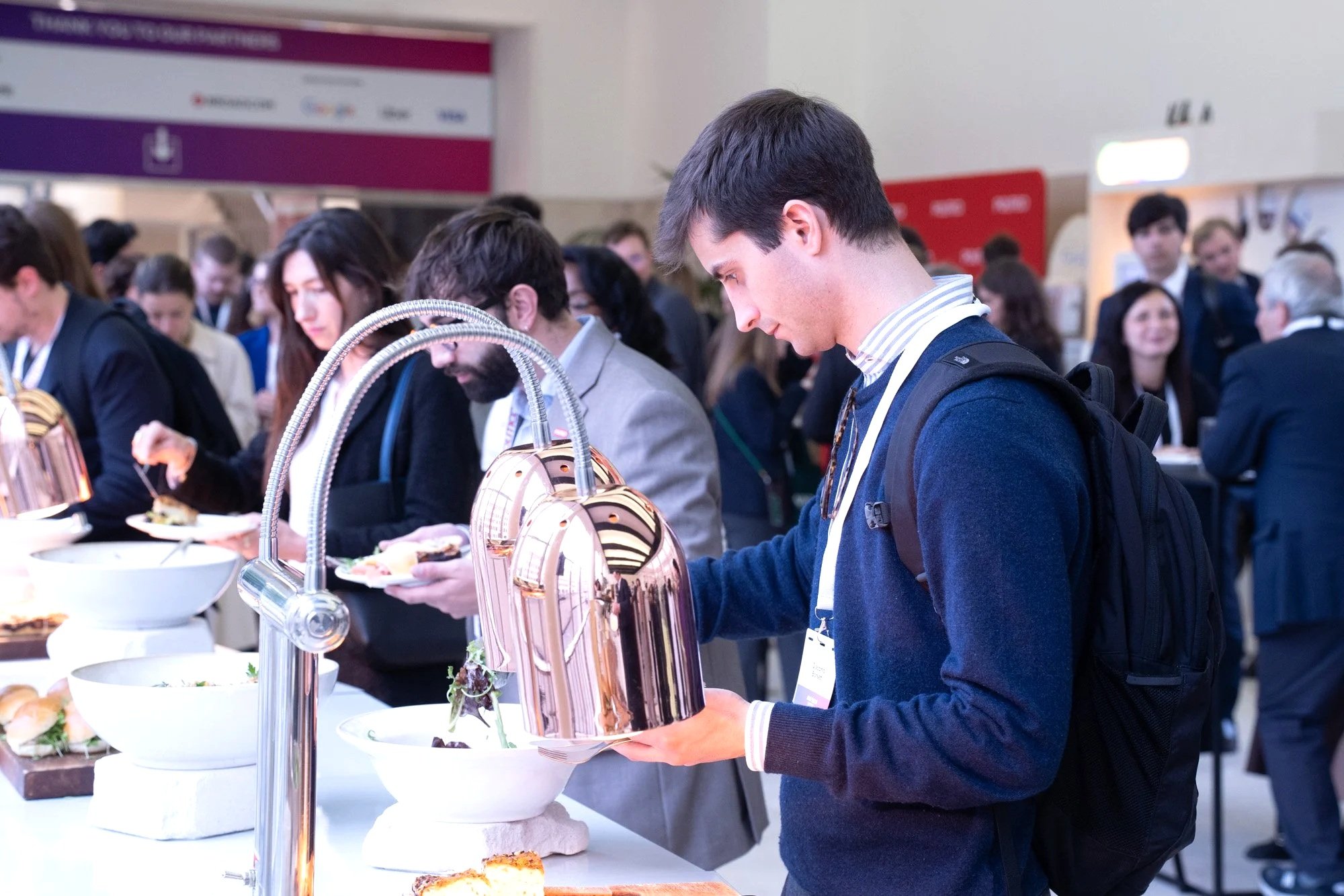A crowd of people at a catering or buffet station, with a young man in the foreground wearing a navy sweater and a backpack, serving himself or examining food from a white platter. There are various dishes and serving utensils on the counter.
