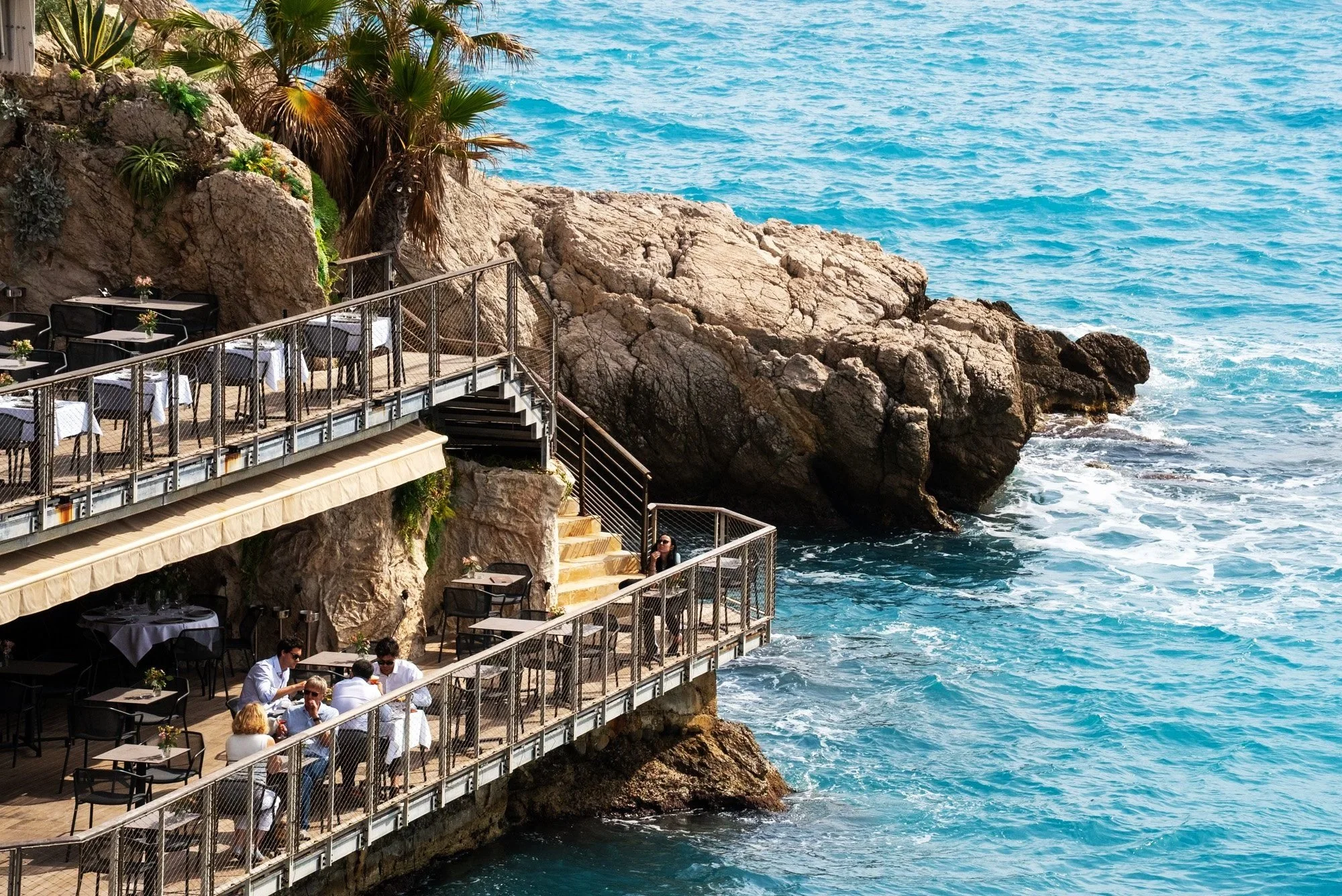 The image shows an outdoor restaurant on a cliffside overlooking the ocean with a rocky coastline and blue water, featuring tables with white tablecloths and people dining. Event Photography Nice, France for Passaporta.eu