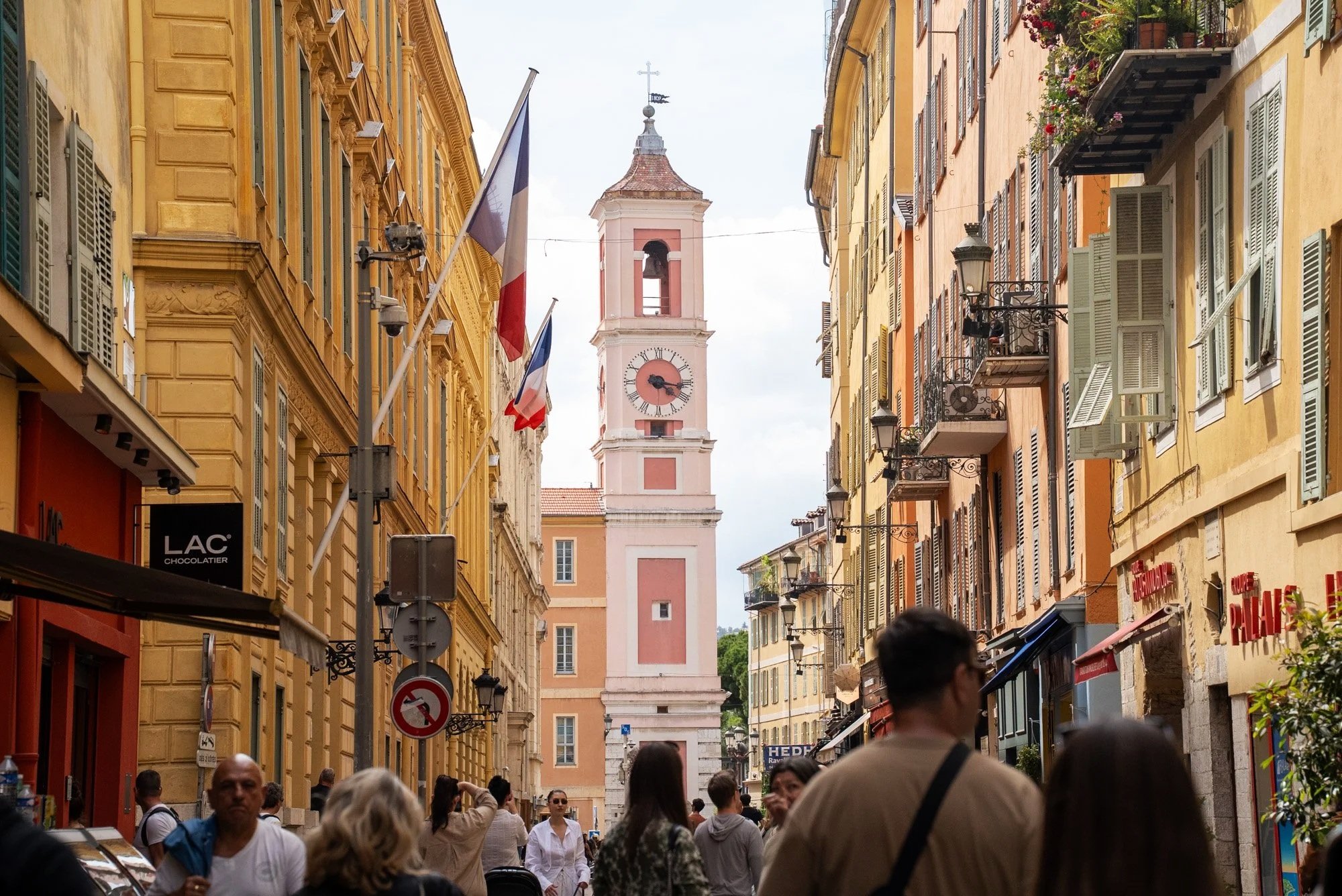 Street scene in a European city with colorful old buildings, flags, a clock tower in the background, and pedestrians walking along the sidewalk.  Event Photography in Monaco for Passaporta.eu