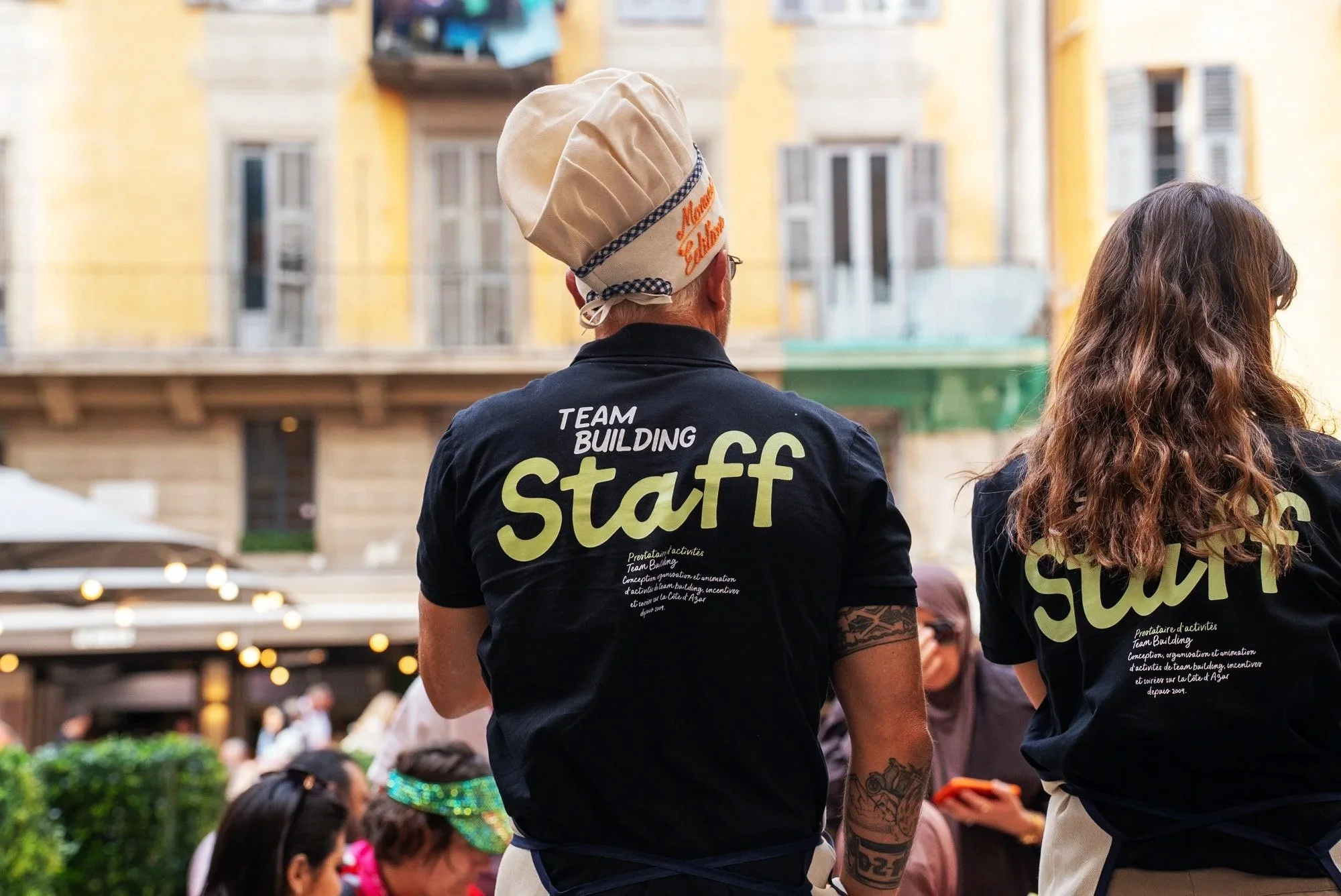 Two event staff members are seen from the back wearing black t-shirts with yellow lettering, one of whom is also wearing a beige chef hat. Event Photography in Monaco for Passaporta.eu