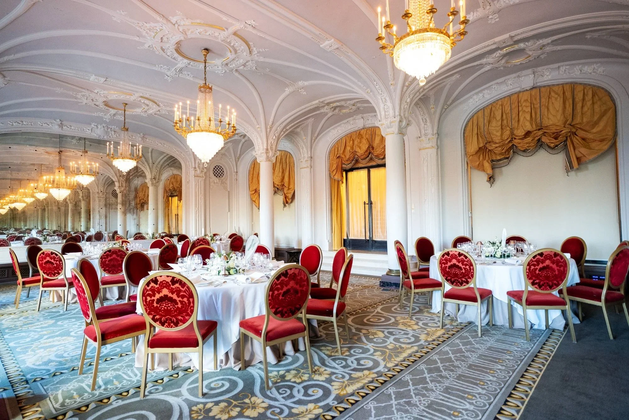Elegant banquet hall with ornate white ceilings, gold chandeliers, and large mirrors. Round tables with white tablecloths, red velvet chairs, and floral centerpieces are set for an event. Event Photography for Passaporta.eu at Hermitage, Monaco