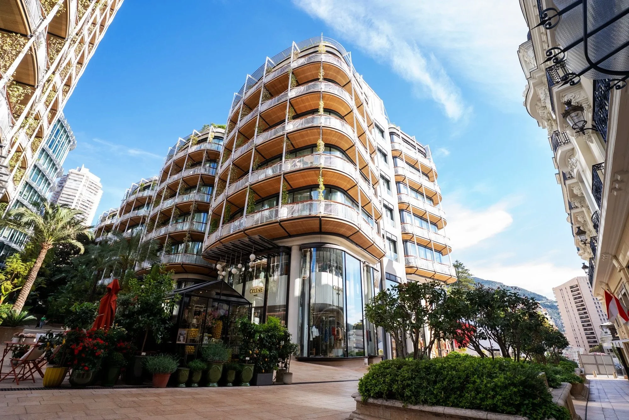 Modern multi-story building with curved balconies and glass storefront, located on a bustling city street with greenery and outdoor seating. Event Photography in Monaco for Passaporta.eu.