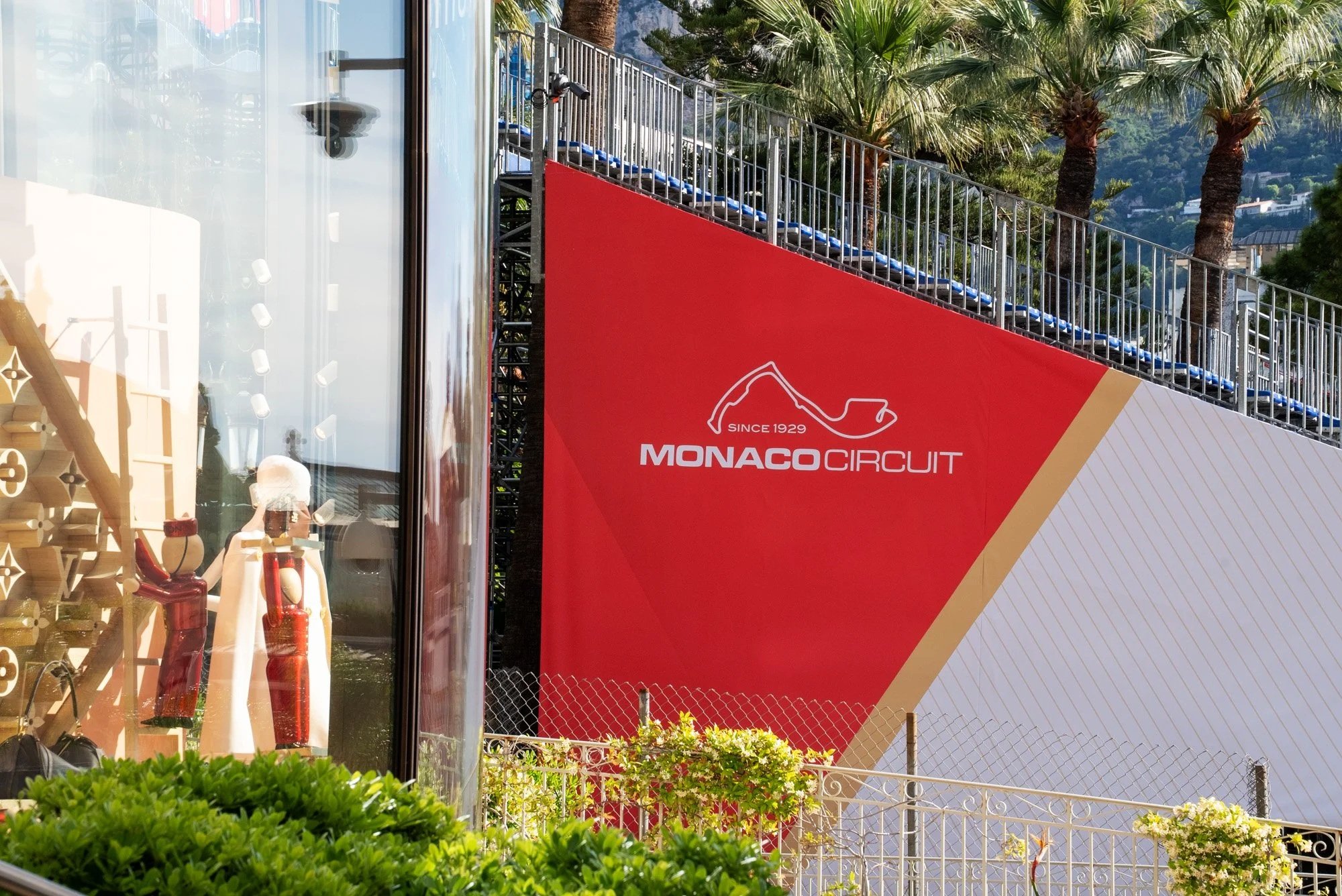 Photo of the Monaco Circuit signage on a red wall with blue grandstand seating above, palm trees in the background, and a view of the racetrack. Event Photography in Monaco for Passaporta.eu