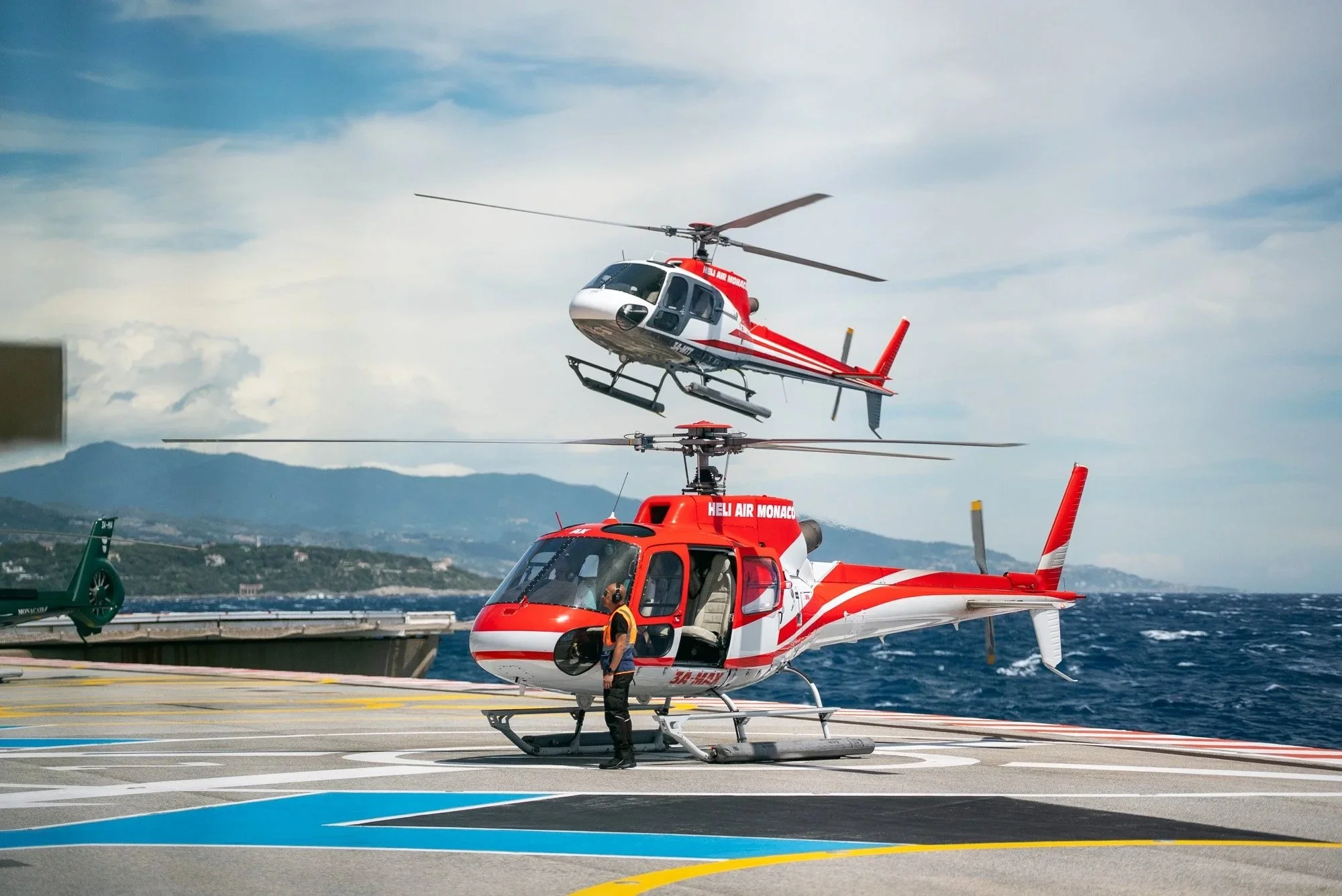 Two helicopter rescue aircrafts on a helipad with a person standing near the front helicopter. The helicopters are marked with red and white colors, with the words "HELI AIR MONACO" visible on one. Event Photography in Monaco for Passaporta.eu.