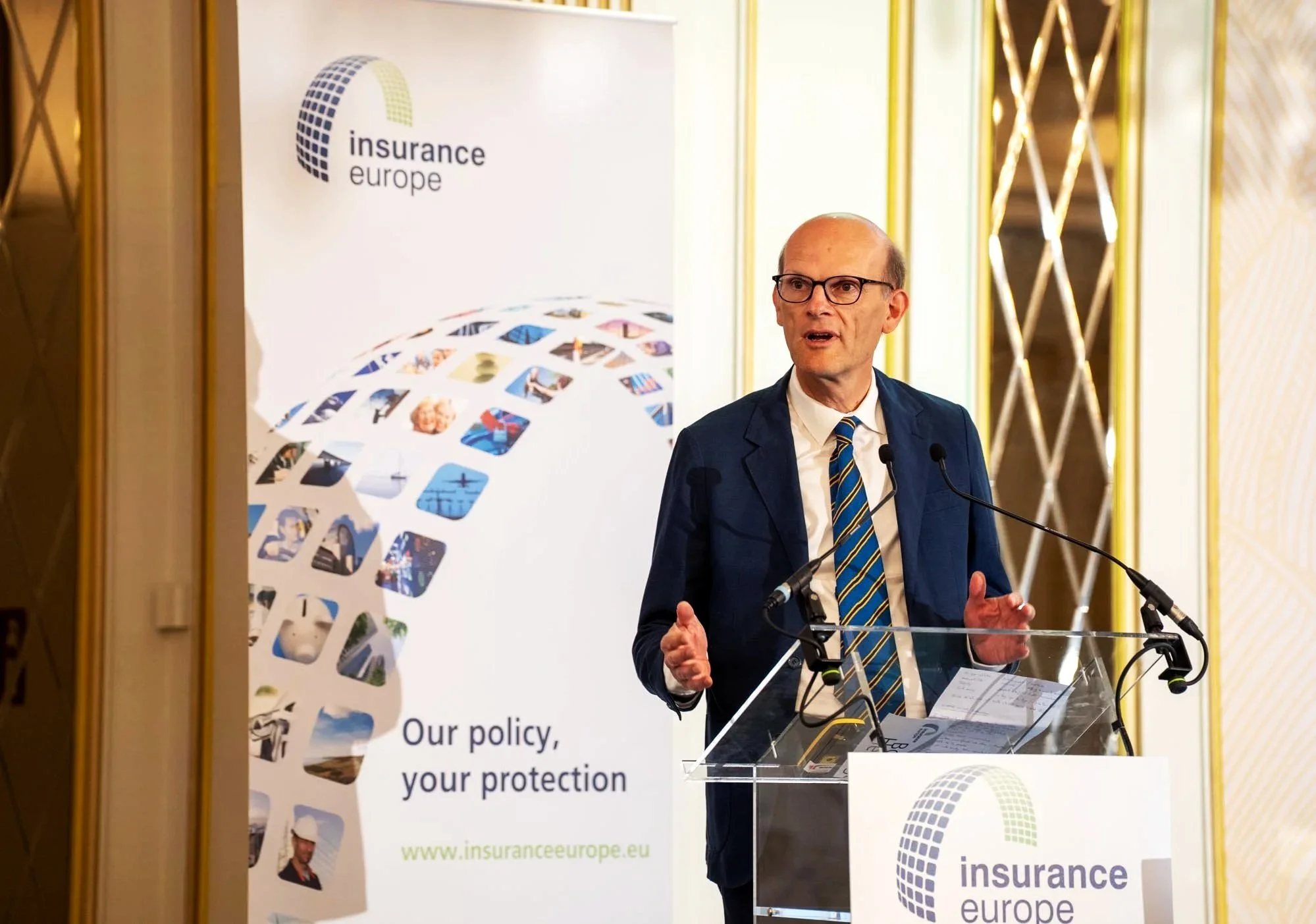 A man in a blue suit, white shirt, and striped tie speaking at a podium with microphones in front of a banner that reads 'Insurance Europe.' The background features decorative golden wall panels, and the banner has a globe logo and images related to 