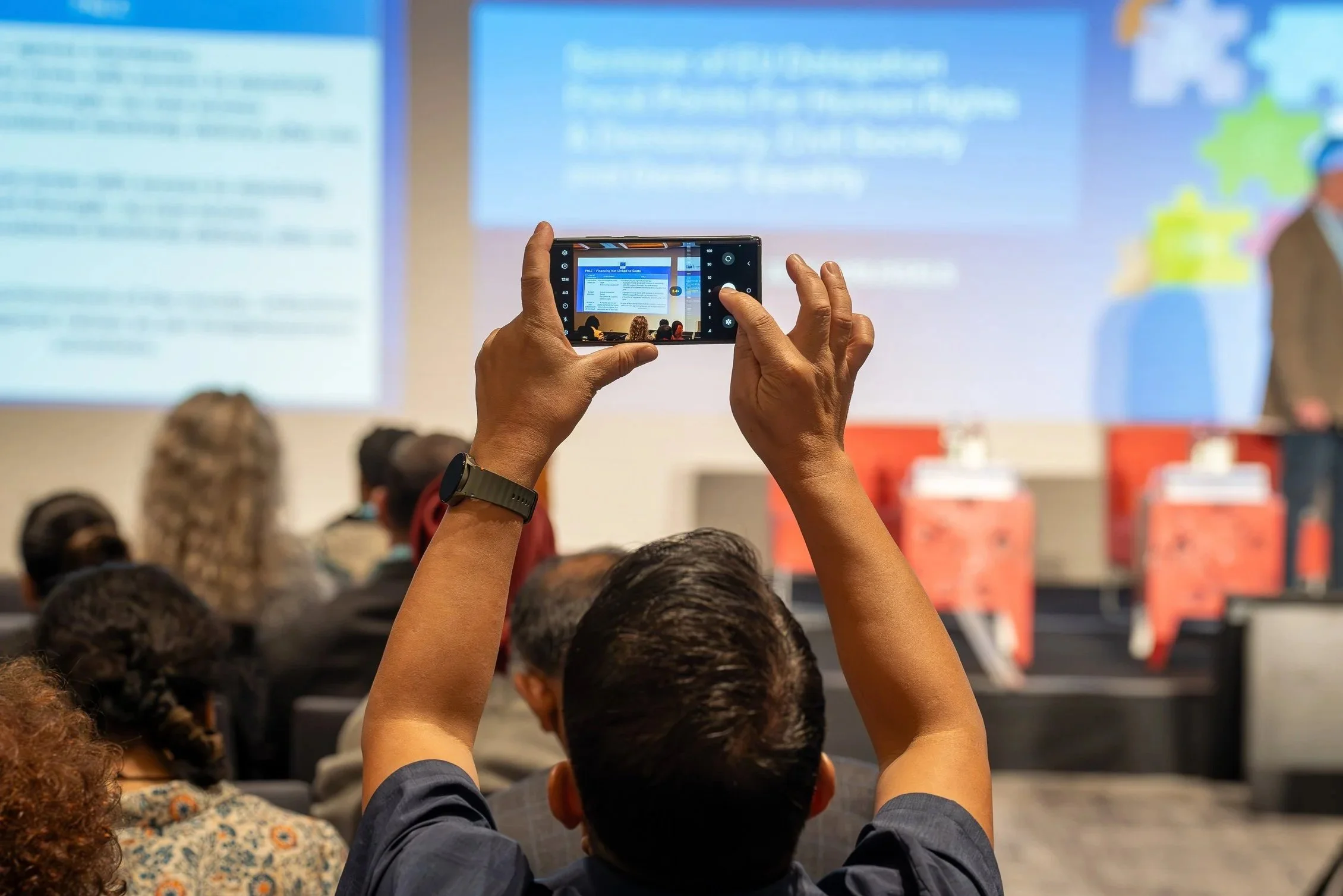 Person holding a smartphone capturing a presentation in a conference room filled with an audience. EU Delegation Seminar on Human Rights 2025, Democracy & Gender Equality Brussels
