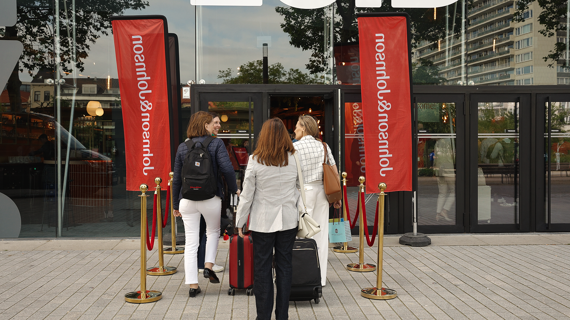 People waiting in line at the entrance of an airport with red flags and gold stanchions, carrying luggage and backpacks. johnson & Johnson Finance day 2026 at Tv studio Play Antwerp Zuid