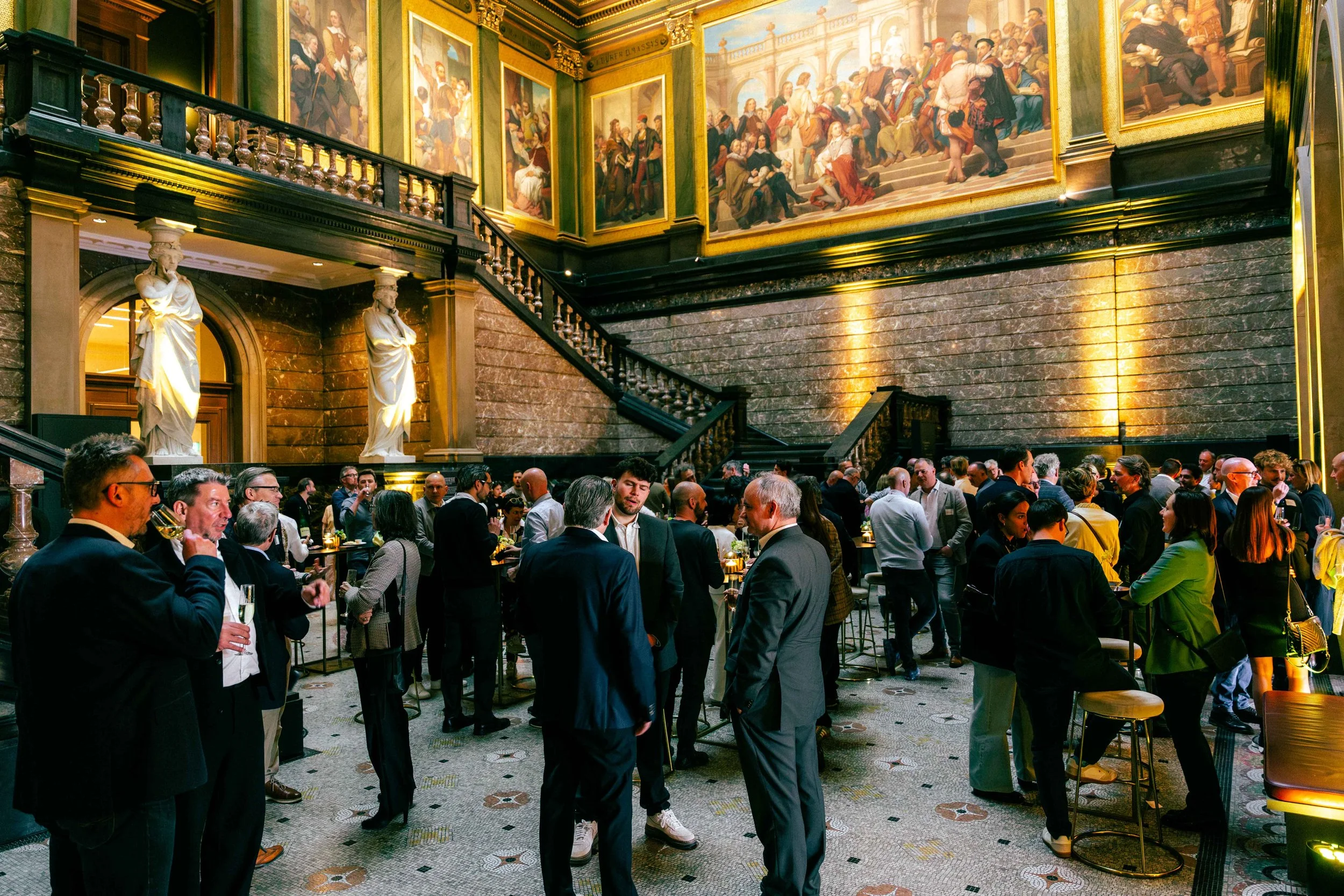 A busy indoor event with many people socializing, dressed in formal attire, in a grand hall with ornate decor, large paintings, statues, and a staircase. Network event at KMSKA for company Nelissen, Bulex, Forbo and SOPREMA.