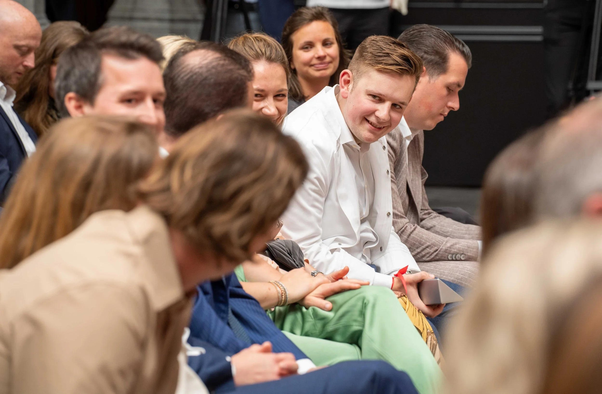 A group of diverse adults sitting in a row, engaged in an event or meeting, some looking down, others looking at the camera with a young man smiling. Michelin guide Ceremony 2026 Antwerp, Handelsbeurs. Star chef