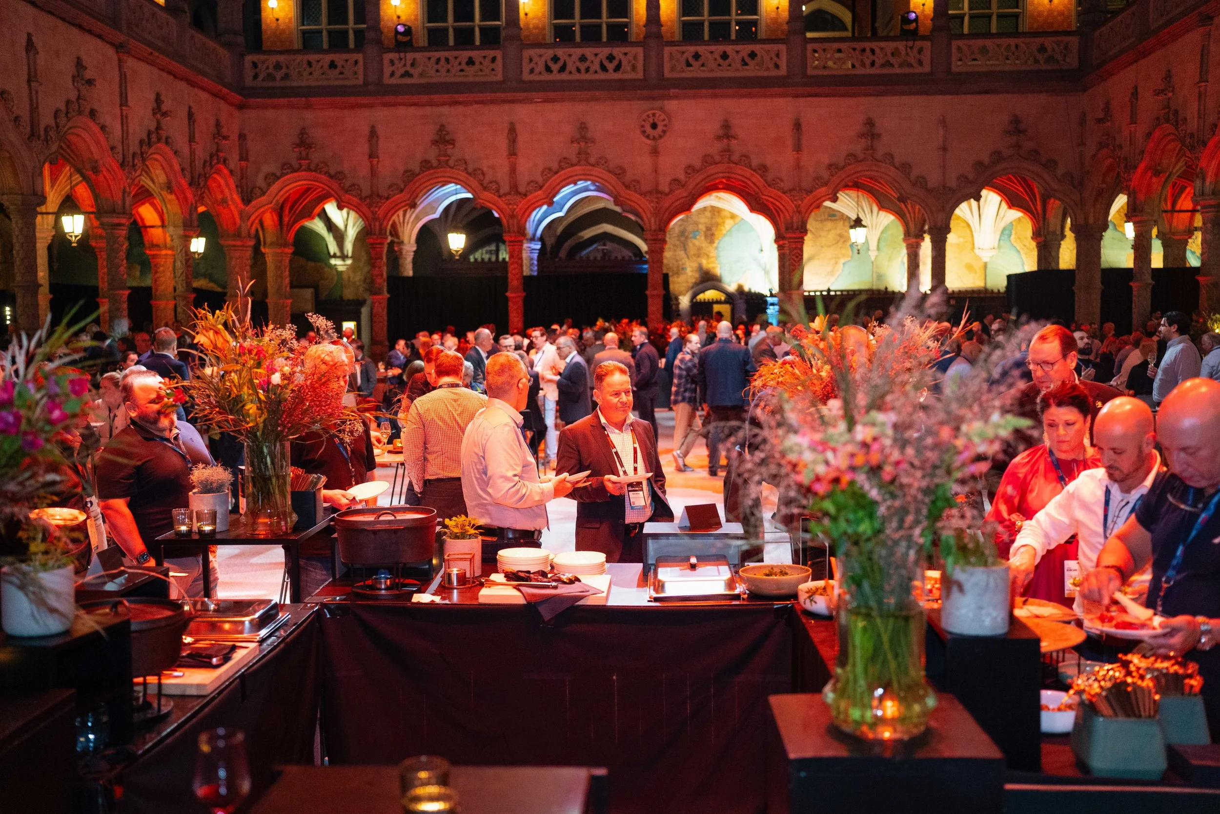A crowded indoor event with many people socializing, some holding drinks, in a historic hall with stained glass windows and ornate arches, decorated with colorful lighting and floral arrangements.