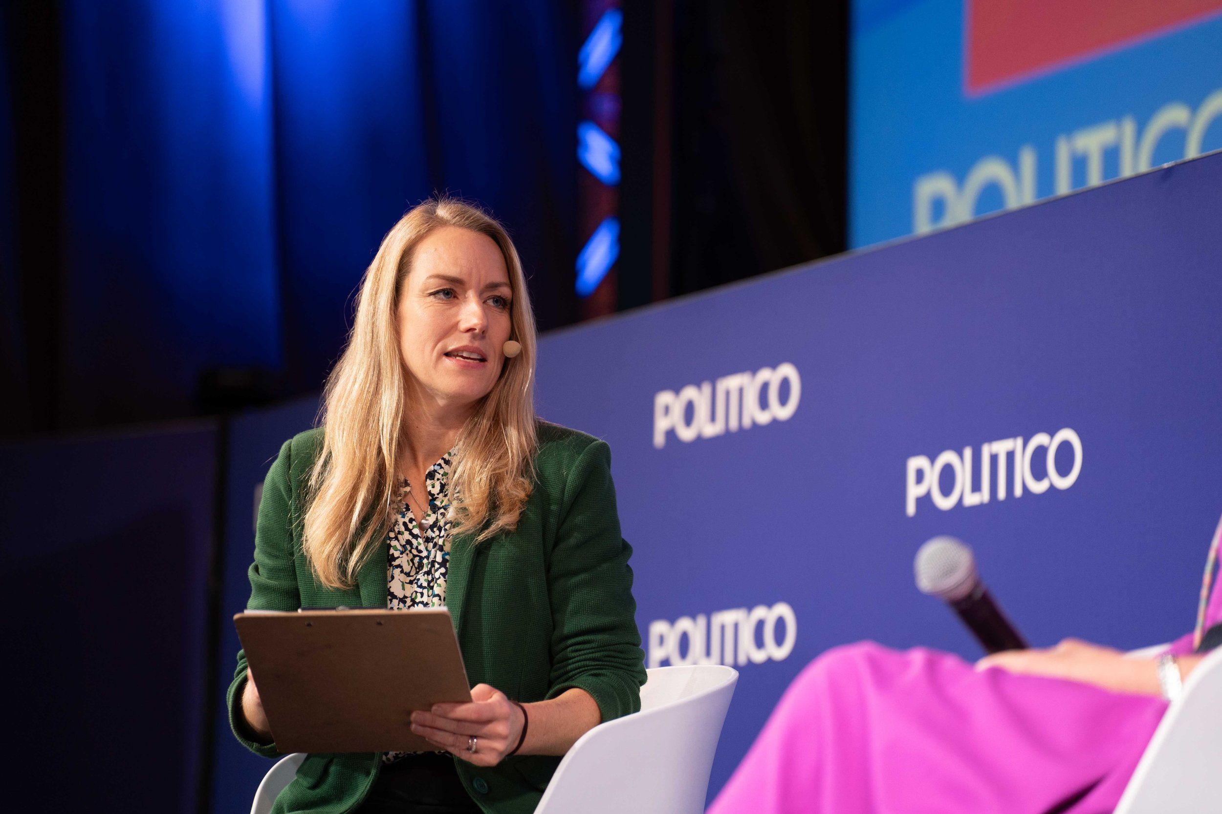 A woman with long blonde hair, wearing a green blazer, sitting on a white chair at a POLITICO event. She is holding a tablet and speaking into a microphone. A large POLITICO banner is behind her. Summit Photographer Brussels.