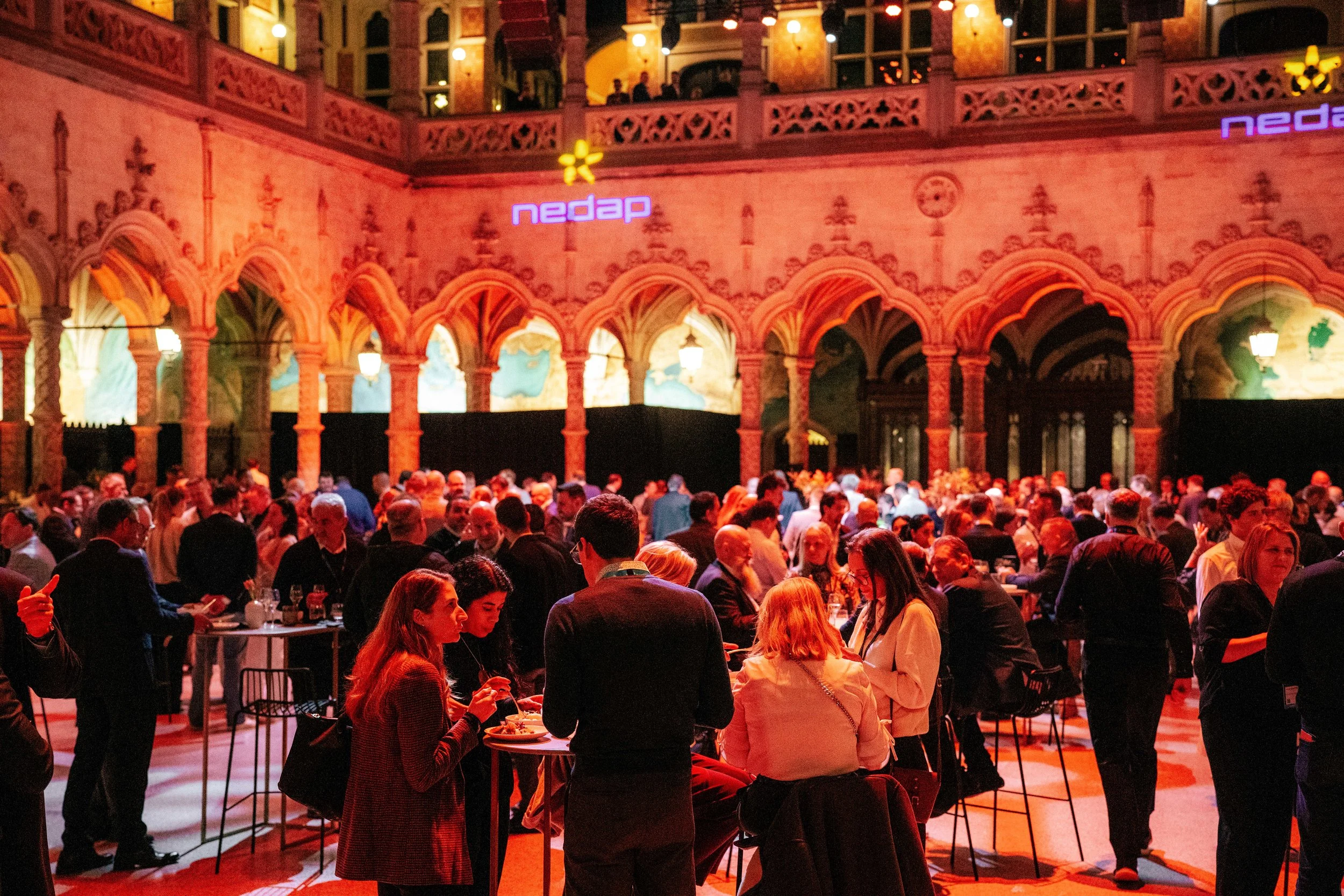 An indoor event with many people seated and standing, in a venue with ornate architecture and colorful lighting, featuring the logo 'nedap' in purple neon lights.