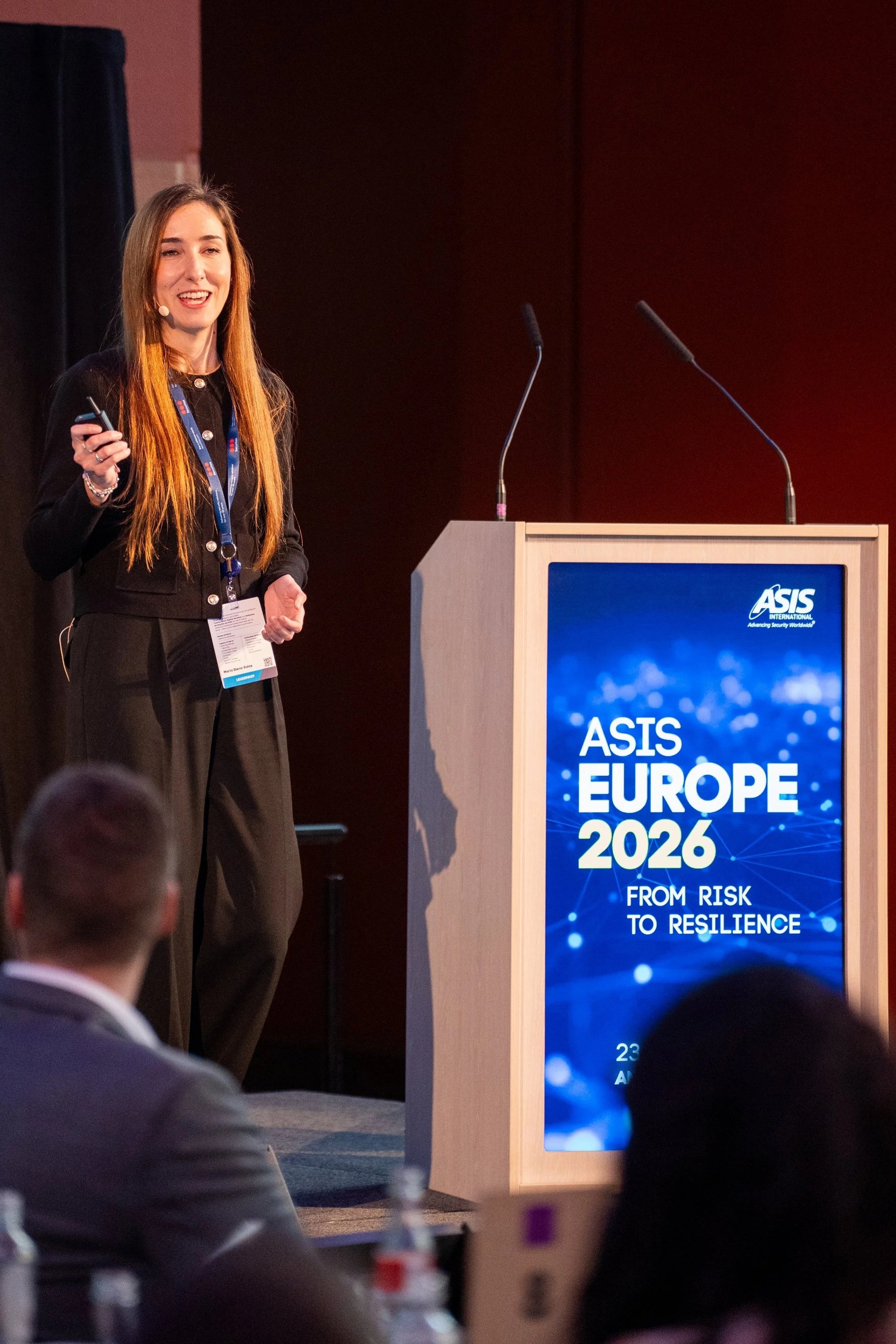 A woman with long hair giving a presentation at a conference, standing next to a podium with a large display screen that reads 'ASIS EUROPE 2026 FROM RISK TO RESILIENCE', with an audience sitting in front.