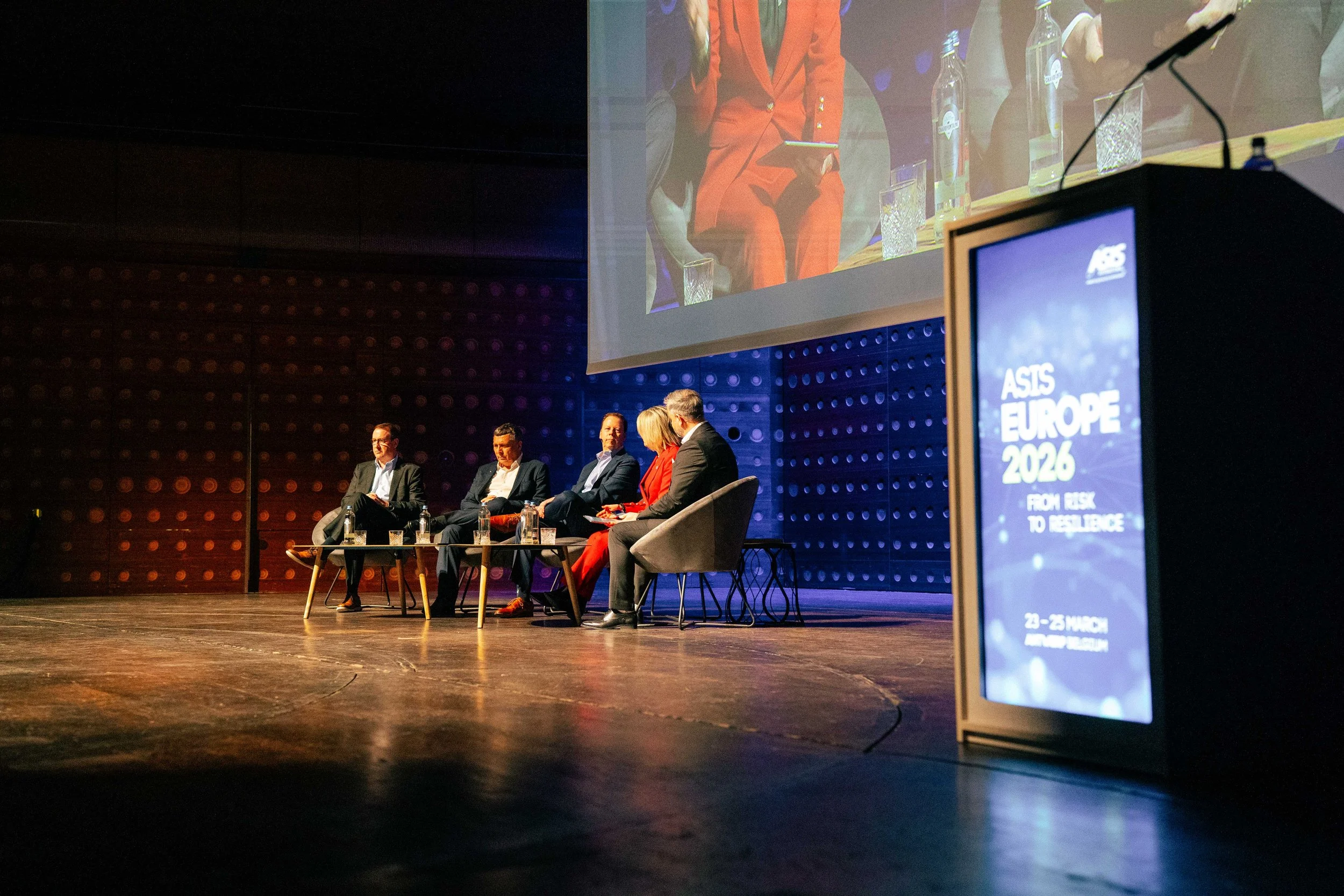 Panel discussion at a conference with five speakers sitting on chairs on a stage, a large screen displaying the panel, and a sign that reads 'ASIS EUROPE 2026.'