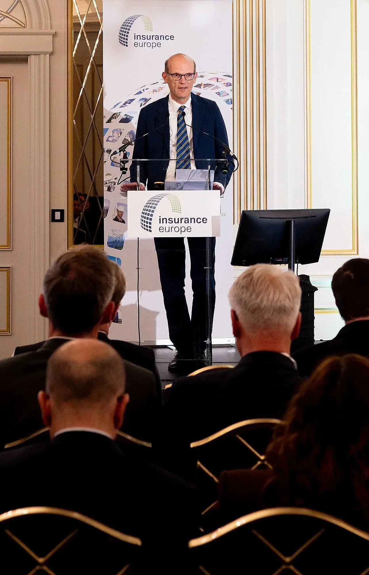 Man standing behind a podium delivering a speech at a conference, with a banner behind him reading 'insurance europe' and an audience seated in front.