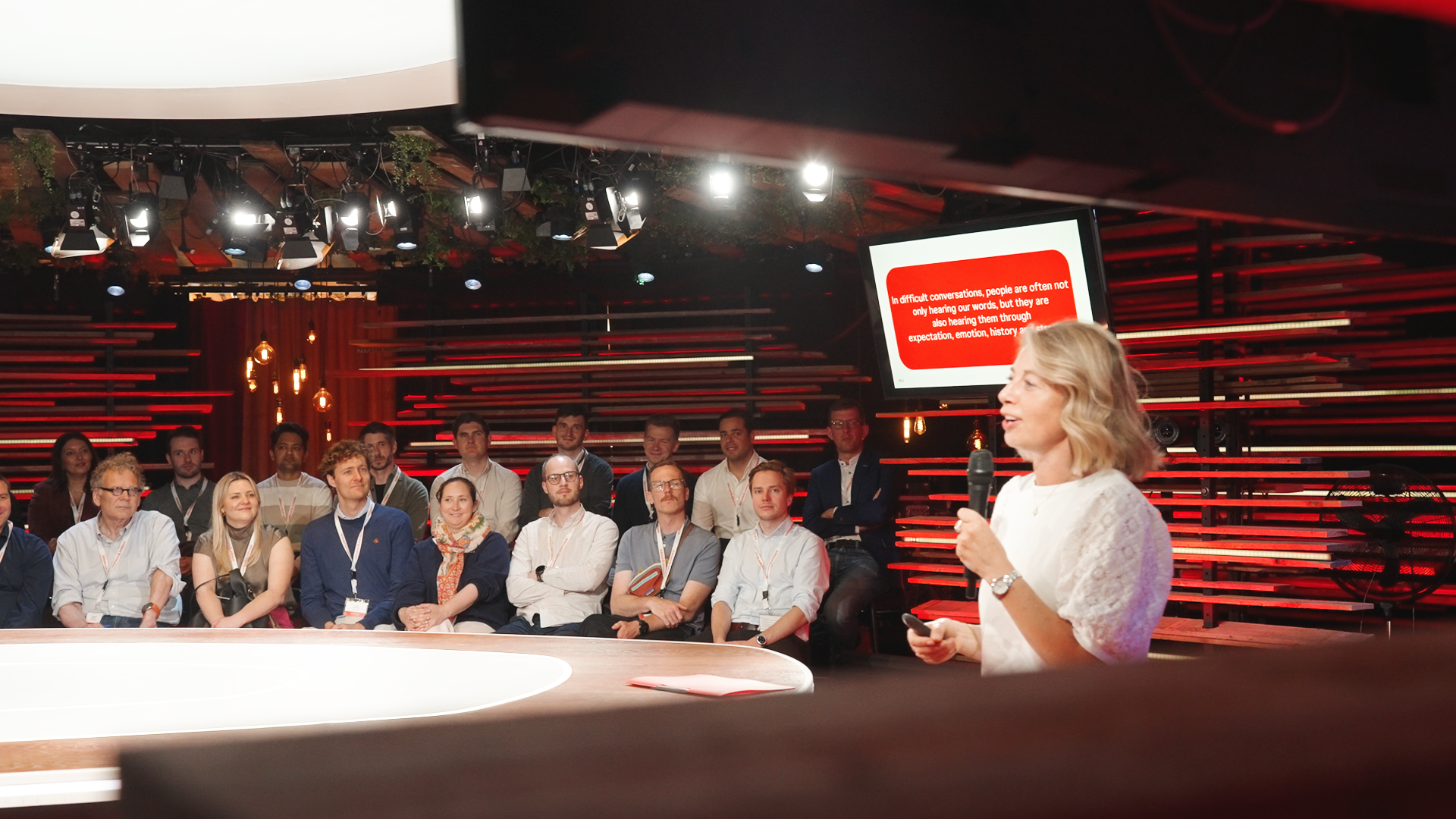 A woman with blonde hair speaking into a microphone in front of a seated audience in a conference setting. A large screen displays a red and white presentation slide behind her. The room has a modern, stylish design with red lighting accents. johnson