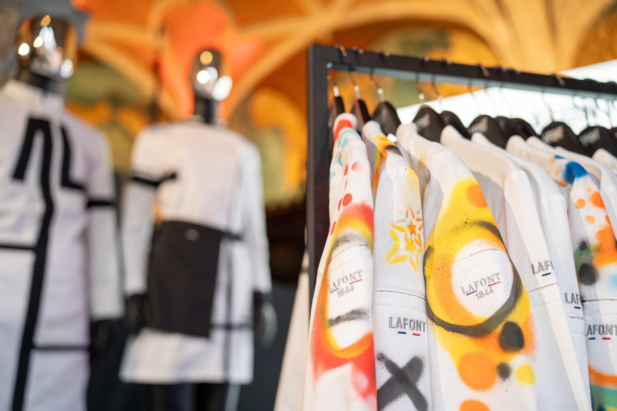 Clothing items hanging on a rack, with colorful abstract spray-painted designs on the fabric, in a retail or display setting. La-font Michelin Guide Ceromony 2026 Antwerp