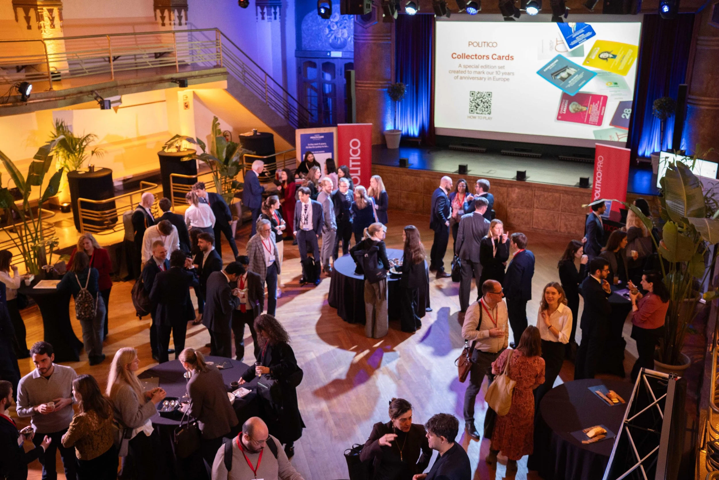 Attendees mingling and networking at a conference or corporate event in a spacious hall with a large screen displaying promotional material and branded banners. Summit Photographer Brussels.
