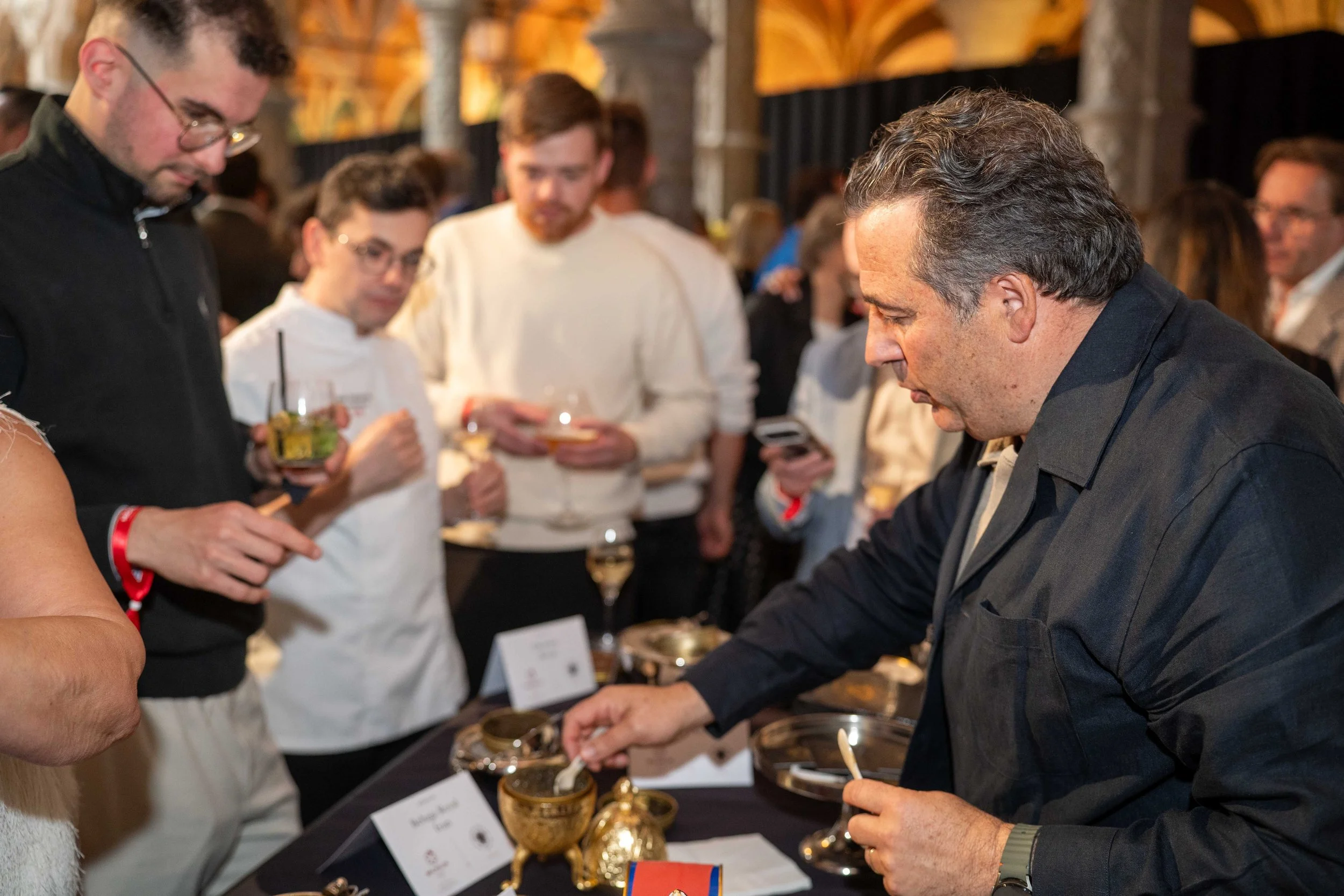 A man in a black suit is at a table with gold-colored ornate objects, possibly antiques or collectibles, while other people in the background are observing and holding drinks. Michelin guide Ceremony 2026 Antwerp, Handelsbeurs. star chefs