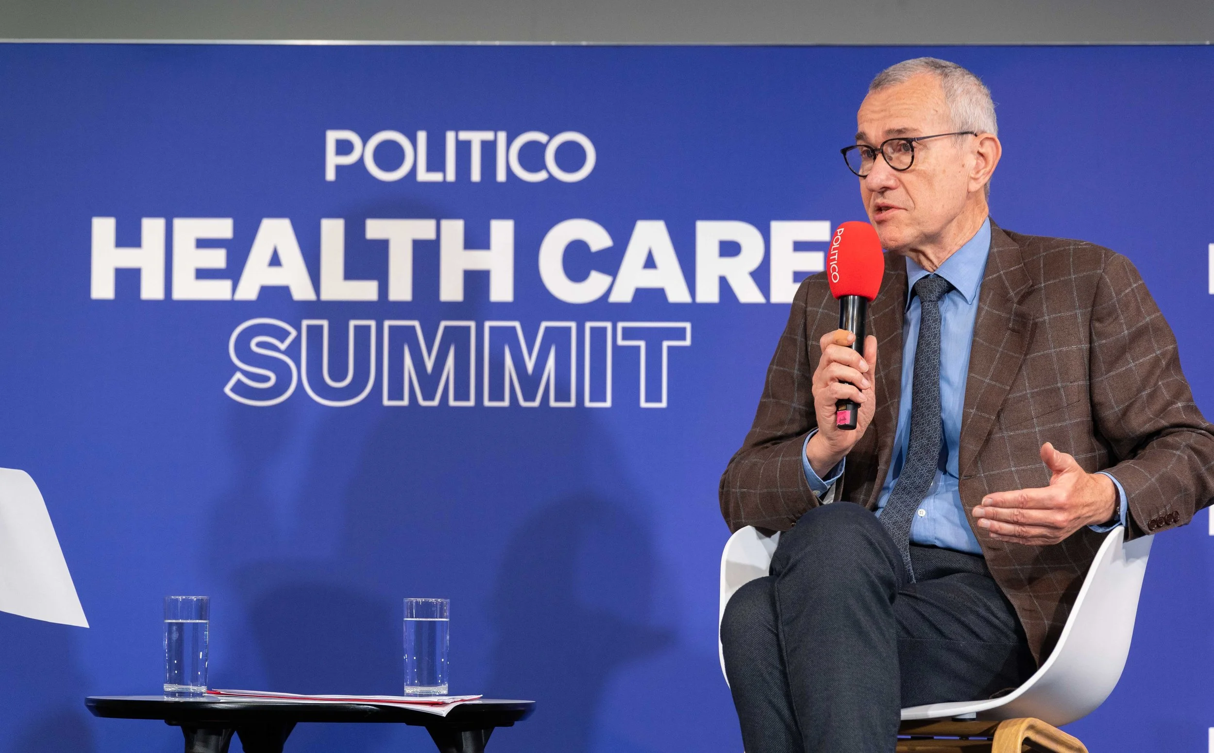 A man wearing glasses and a plaid brown blazer speaking into a red microphone labeled 'POLITICO' at a healthcare summit, sitting on a white chair with a small black table and two glasses of water in front of him. Summit Photographer Brussels.