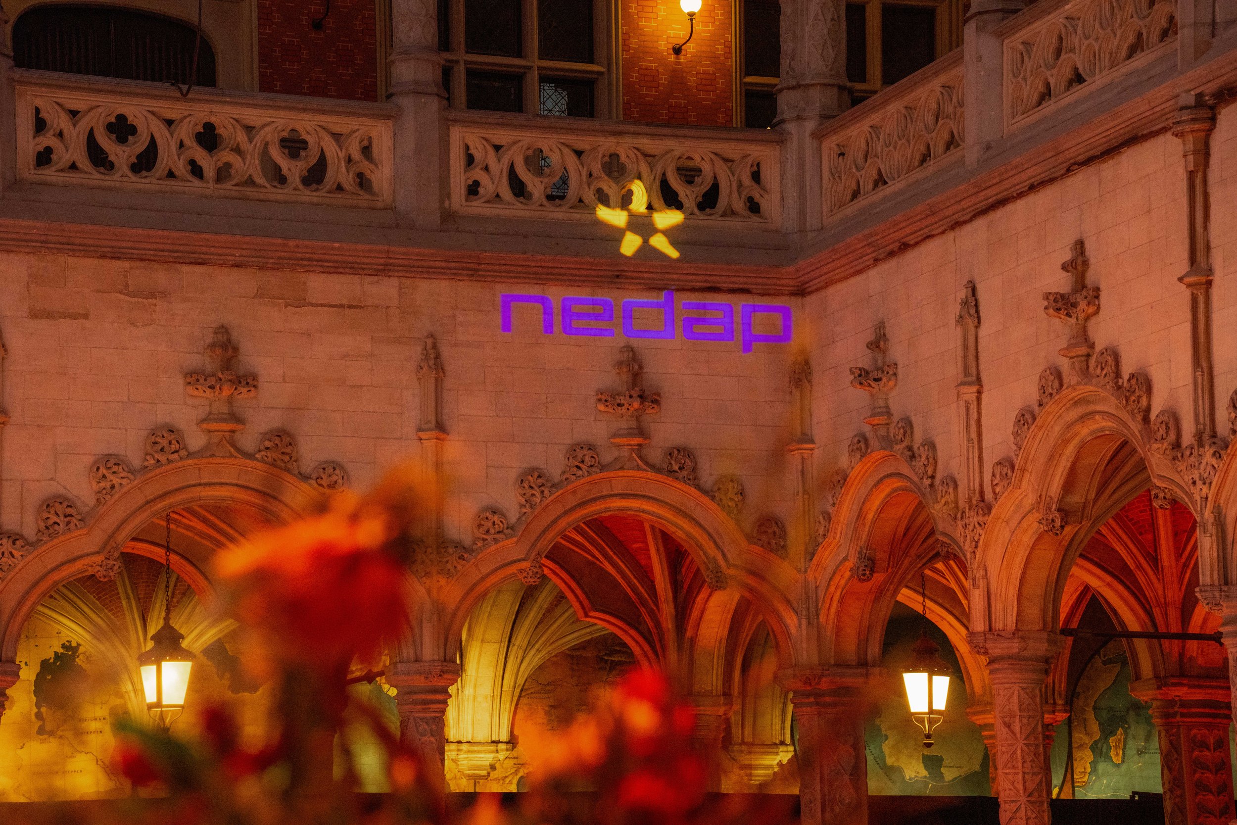 Night view of an ornate building with Gothic architectural style, featuring arched windows and decorative stonework. There are illuminated street lamps and a neon sign that says 'Nedap' in purple, along with a yellow logo of a bee or insect above it.