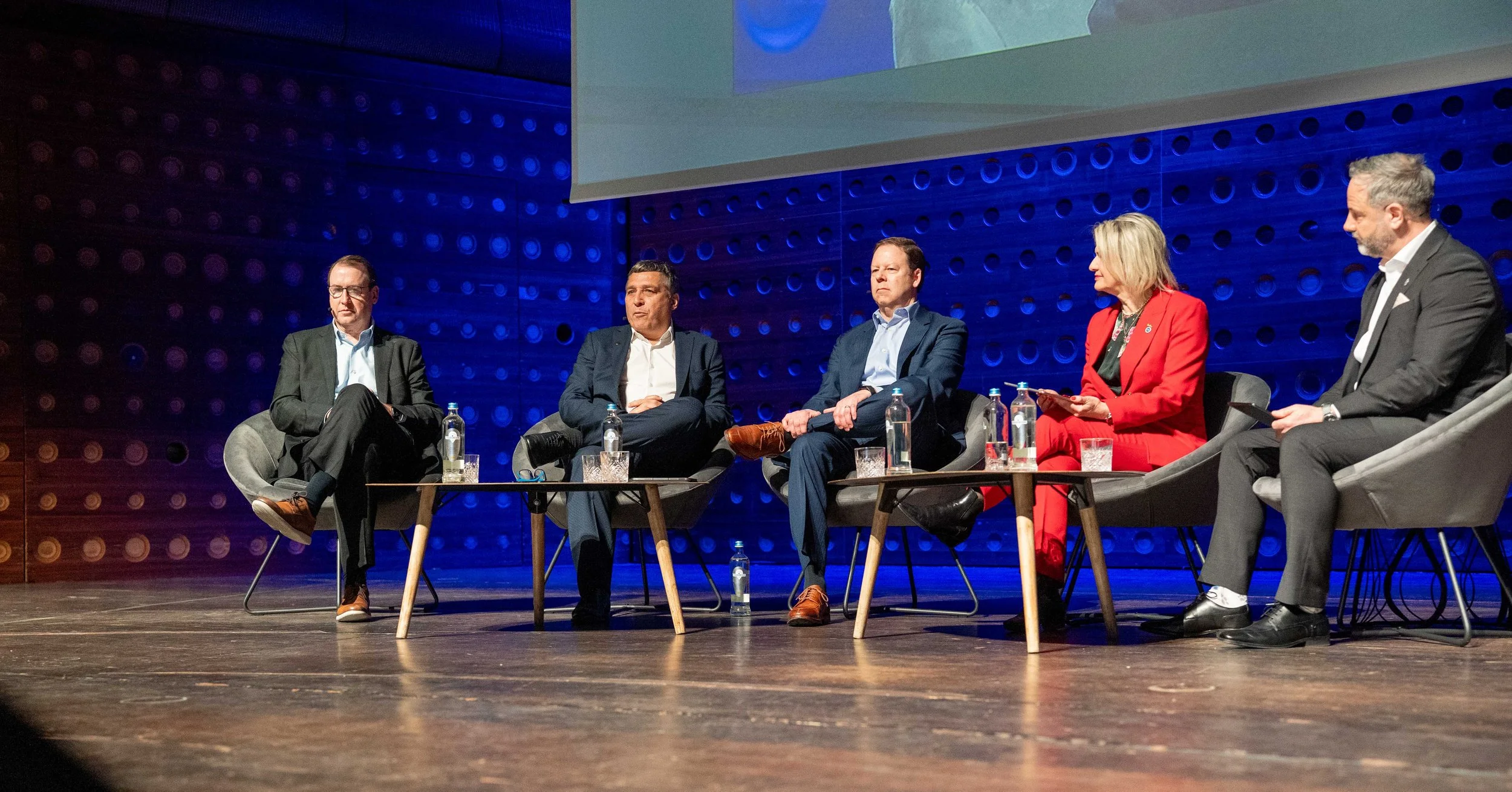 A panel of six people sitting on a stage participating in a discussion, with bottled water and glasses on small tables in front of them. ASIS EUROPE ANTWERP 2026 at the ZOO