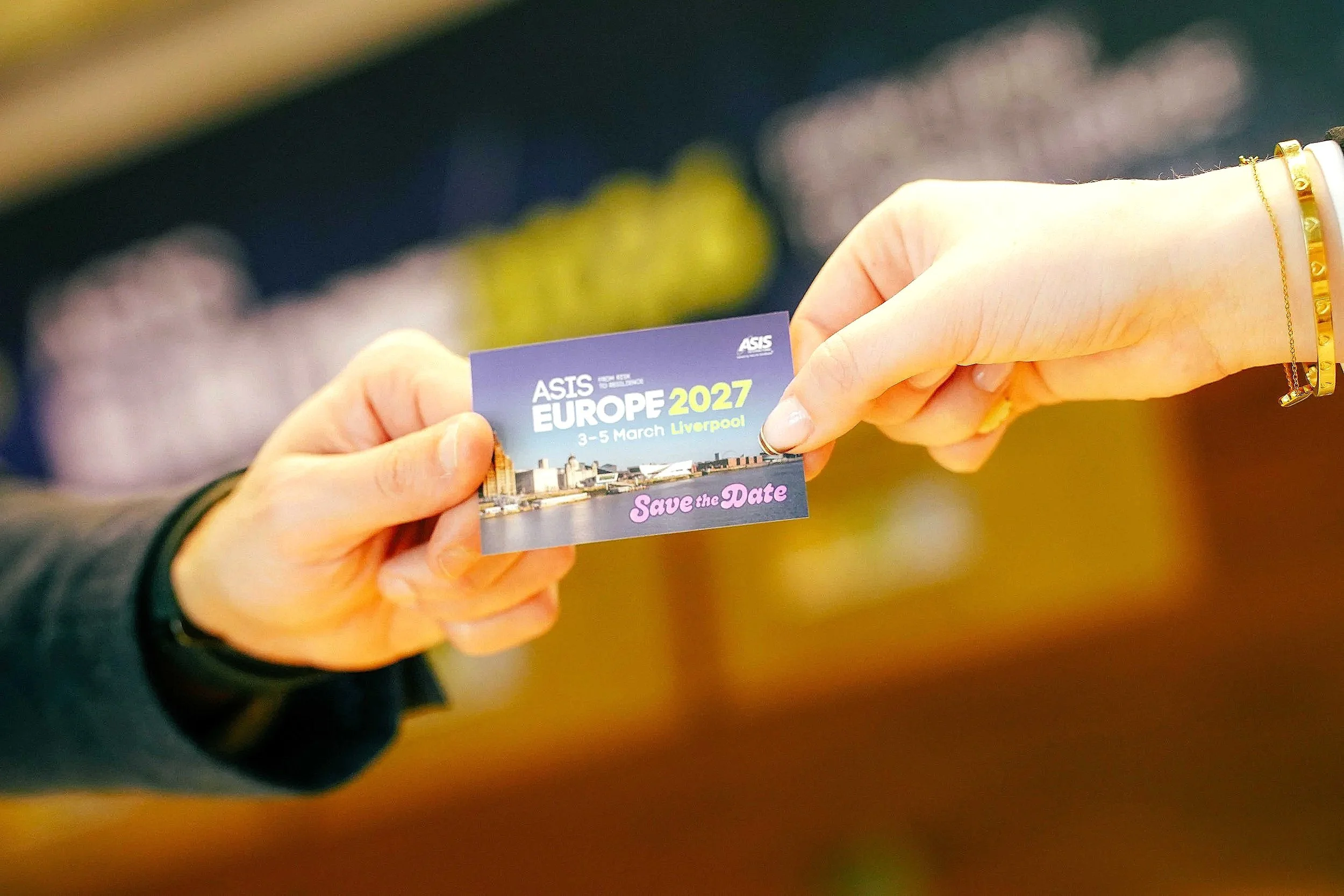Close-up of two hands holding a business card for an event called 'ASIS EUROPE 2027' taking place in Liverpool from March 3-5, with a background of blurred colorful objects.