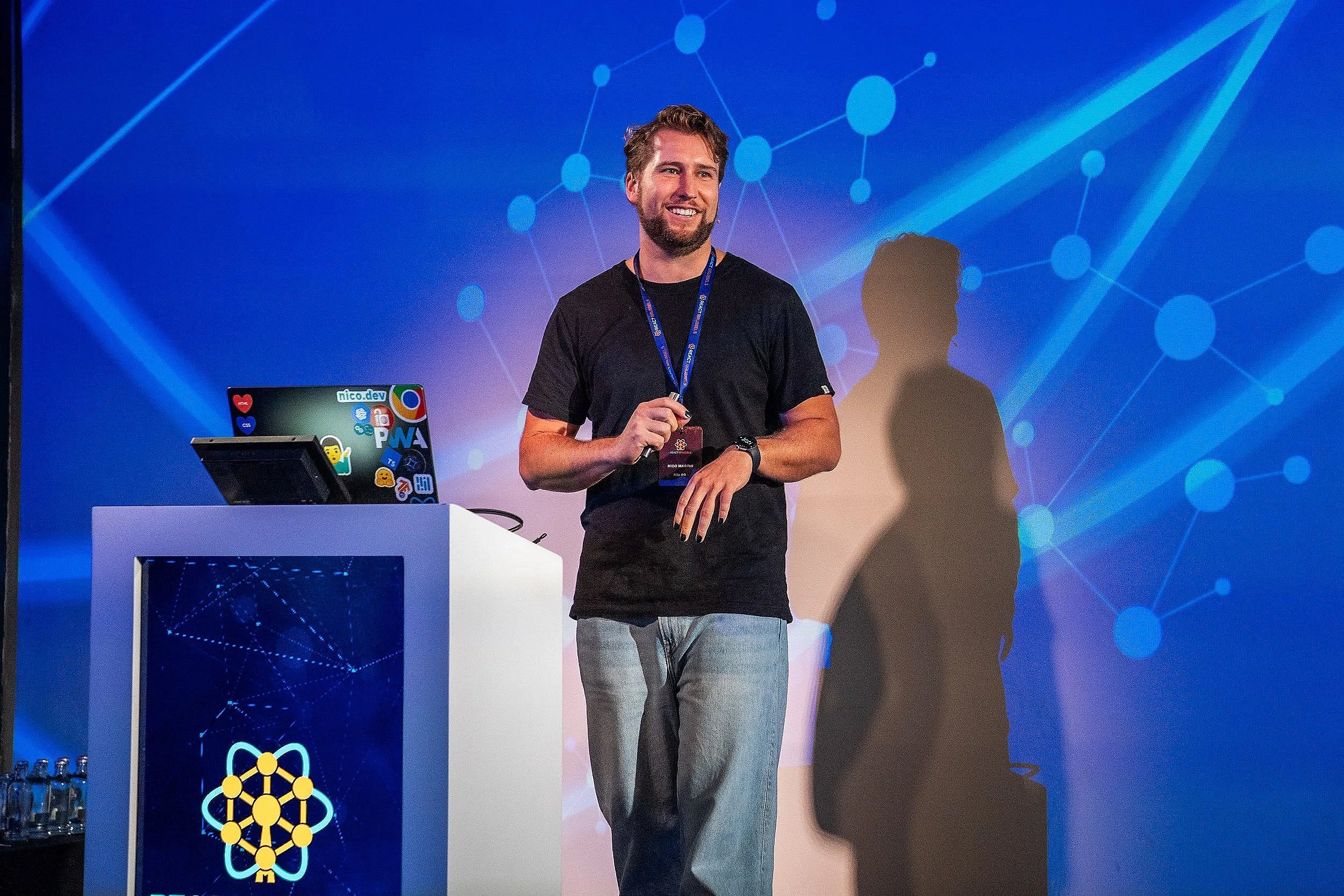 A man with a beard and casual clothing standing on a stage, holding a microphone, giving a presentation with a blue technological background and a podium featuring a network logo.
