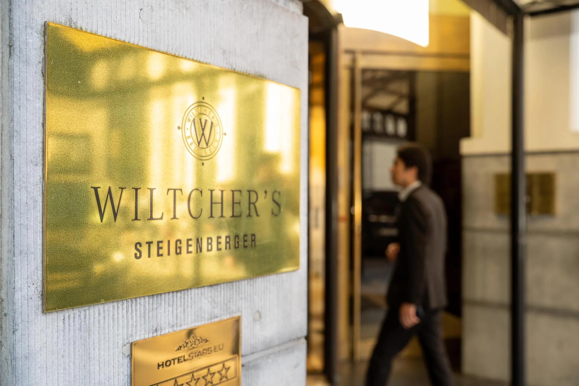 Close-up of a gold hotel sign with the words "Witcher's" and "Steigenberger" visible, mounted on a concrete wall. In the background, a man in a suit is walking past the entrance of the hotel. Insurance Europe’s 16th International Conference at the St