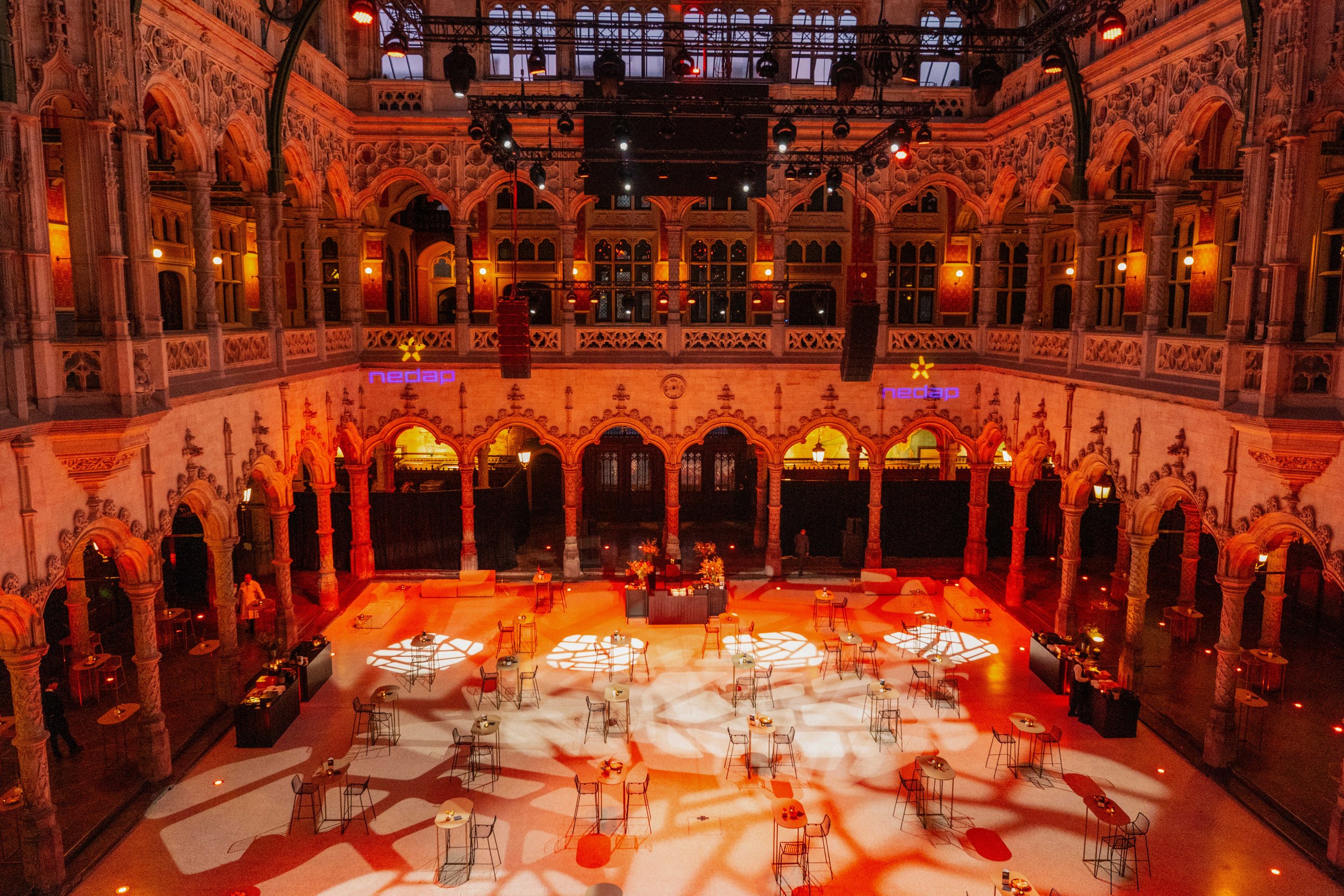 Interior of a large, ornate hall decorated with arches and intricate architectural details, illuminated with warm lighting, with a stage set up with tables and chairs for an event.