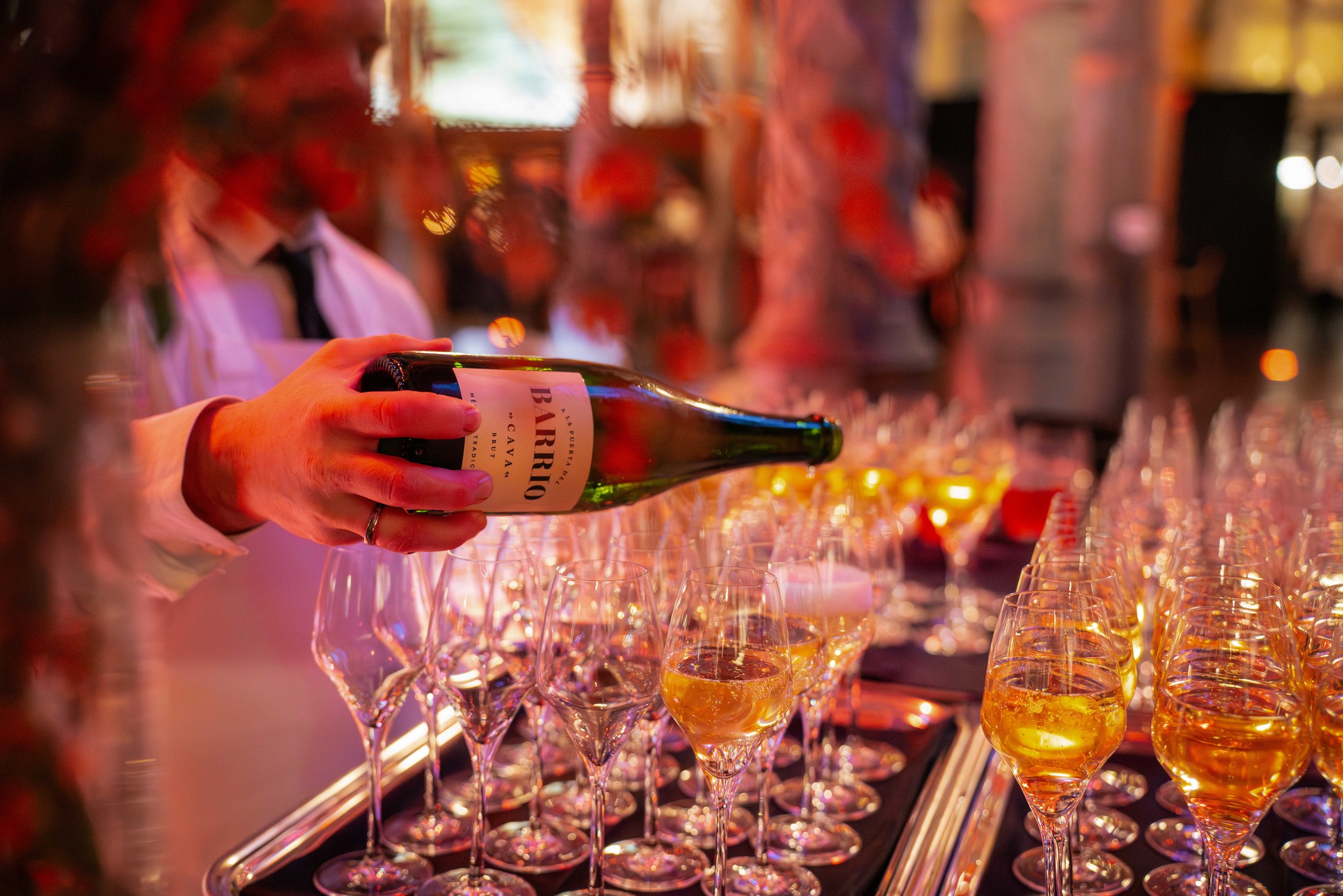 A bartender in a white shirt and black tie pours a drink into a glass at a bar with rows of glasses filled with golden liquid and illuminated by colorful ambient lighting.