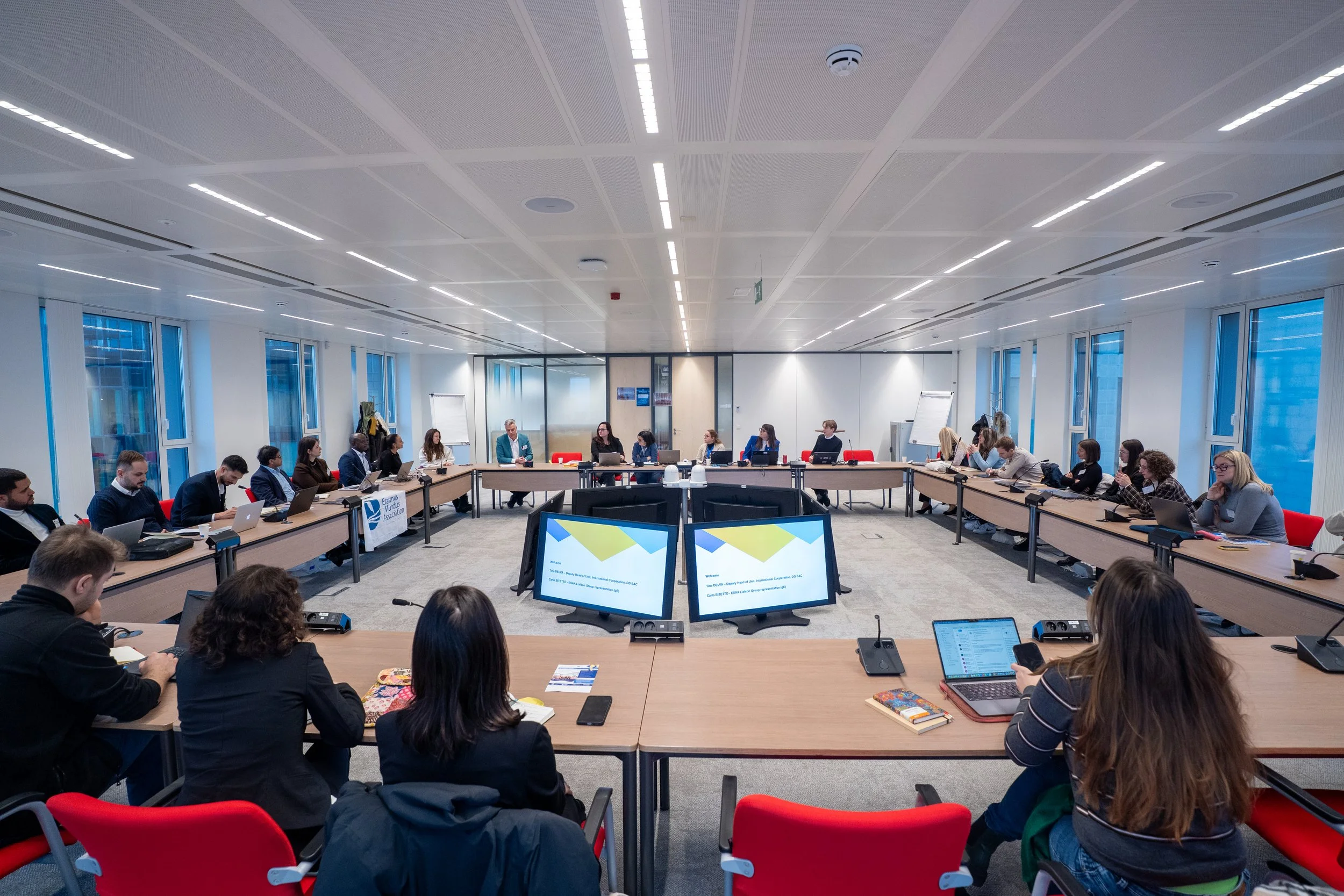 A large conference room filled with people seated around a U-shaped table, attending a meeting or conference. Students of the Erasmus+ ESSA European Comission meeting in Brussels.