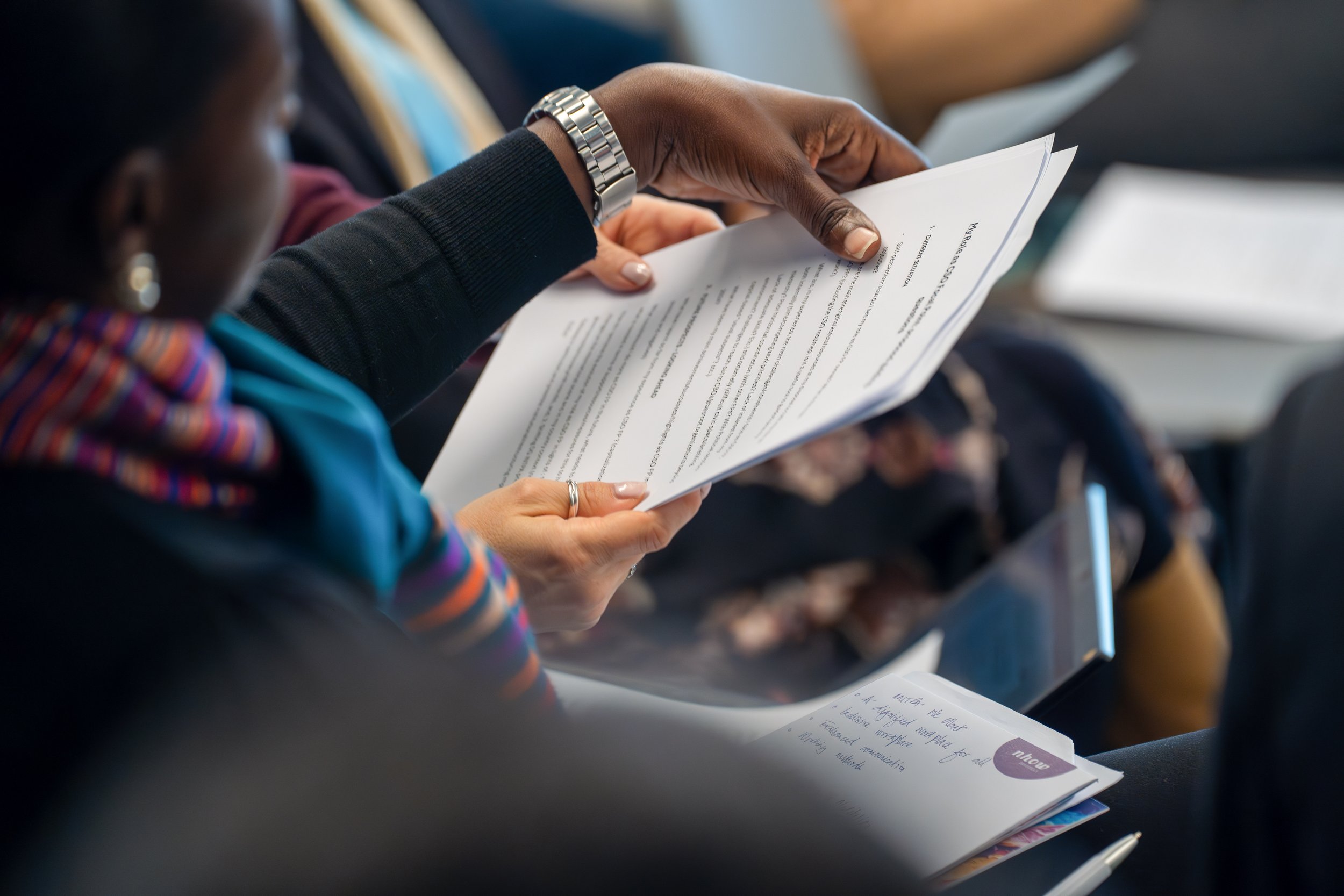 A person with dark skin wearing a silver watch and a black long-sleeve shirt reading a printed document or pamphlet at a conference or seminar. EU Delegation Seminar on Human Rights 2025, Democracy & Gender Equality Brussels