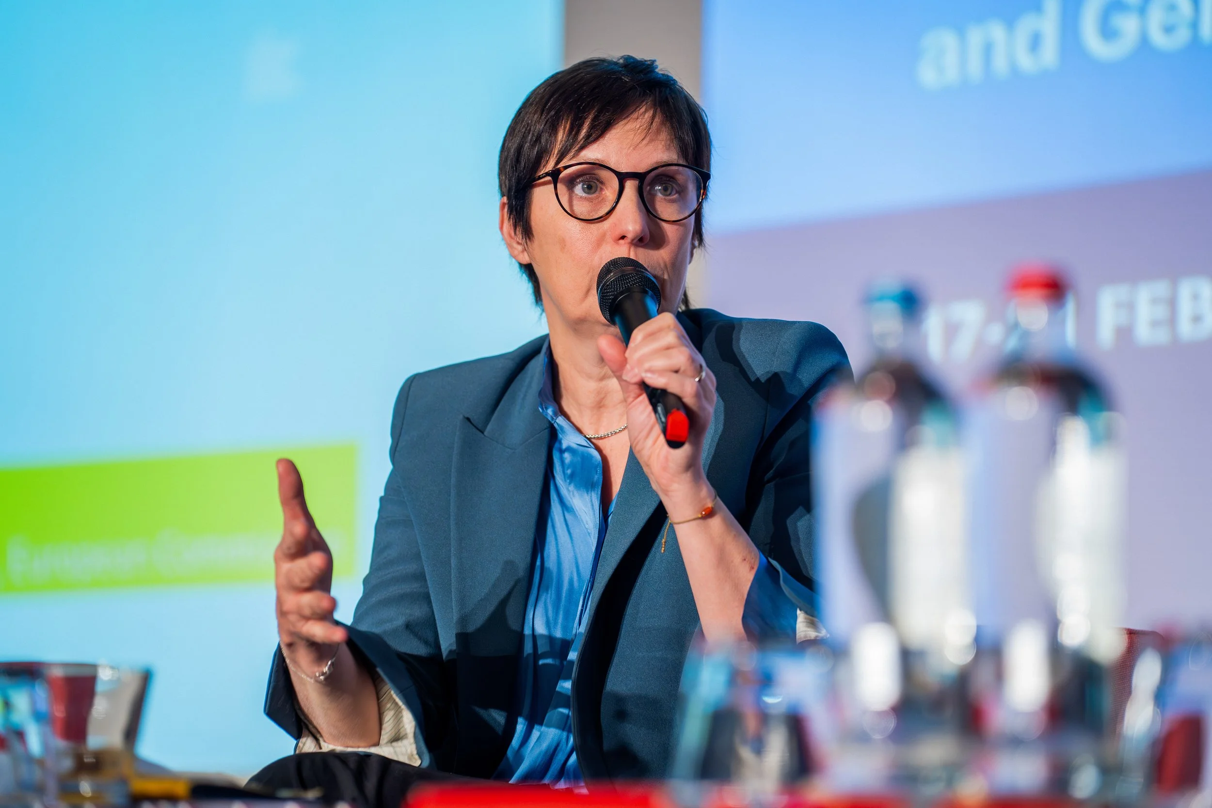Woman with short dark hair and glasses, speaking into a microphone at a conference or presentation. EU Delegation Seminar on Human Rights 2025, Democracy & Gender Equality Brussels