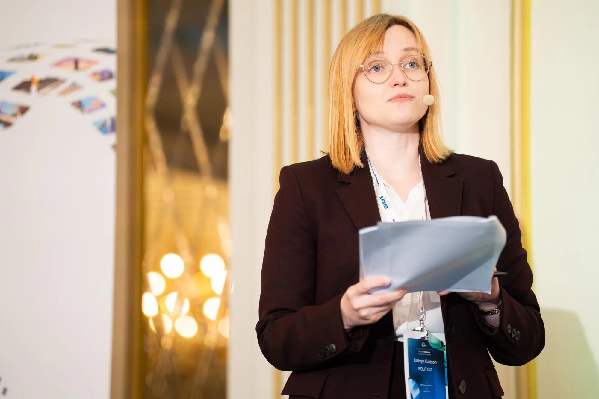 A woman with red hair, glasses, and a dark blazer is standing indoors, holding a folder and reading. She is wearing a conference badge. The background features decorative lighting and a blurred wall. Insurance Europe’s 16th International Conference