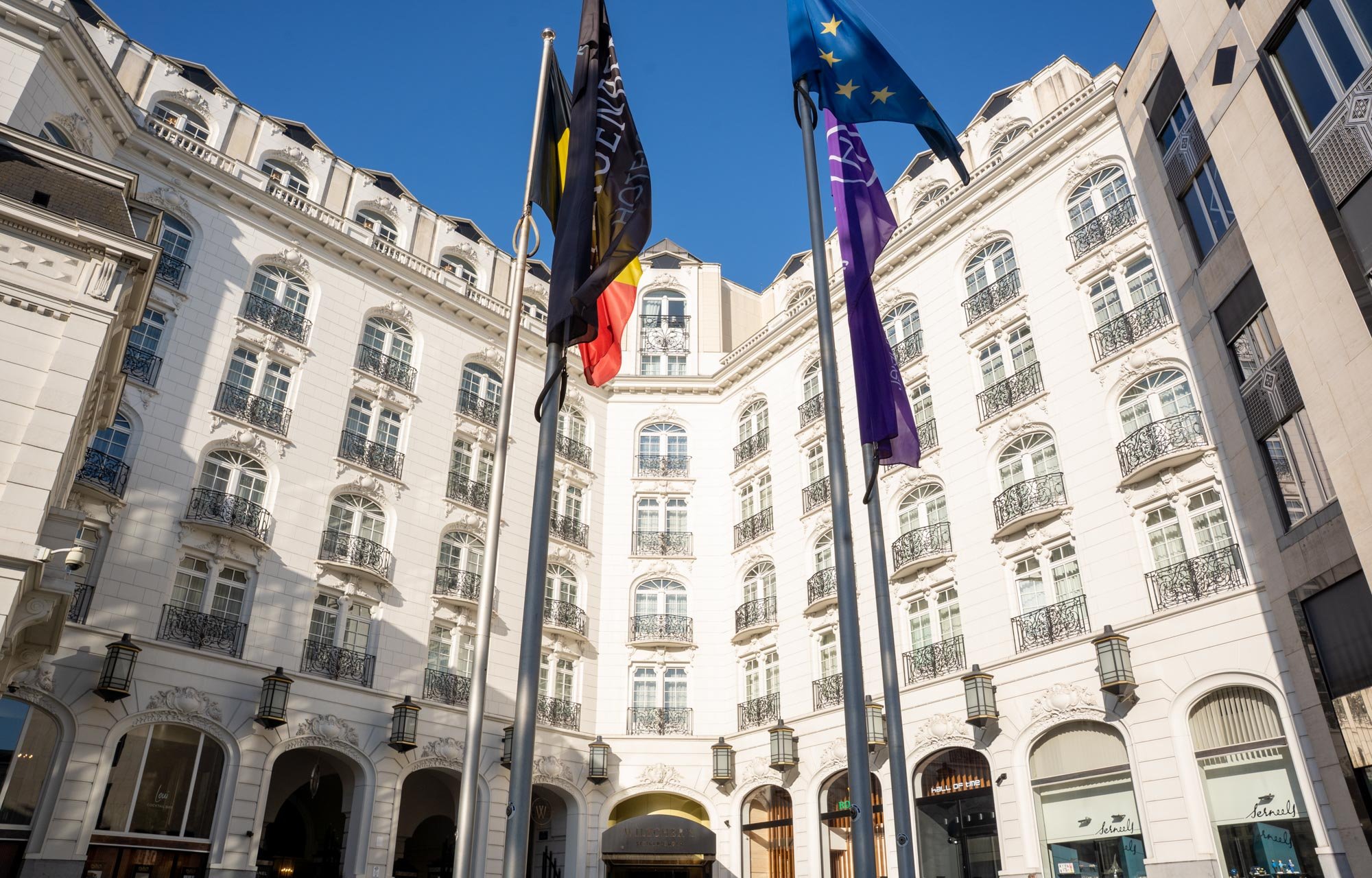Multiple flags flying in front of a white ornate building with balconies and arched windows, under a clear blue sky. Insurance Europe’s 16th International Conference at the Steigenberger Icon Wiltcher’s Hotel, Brussels.