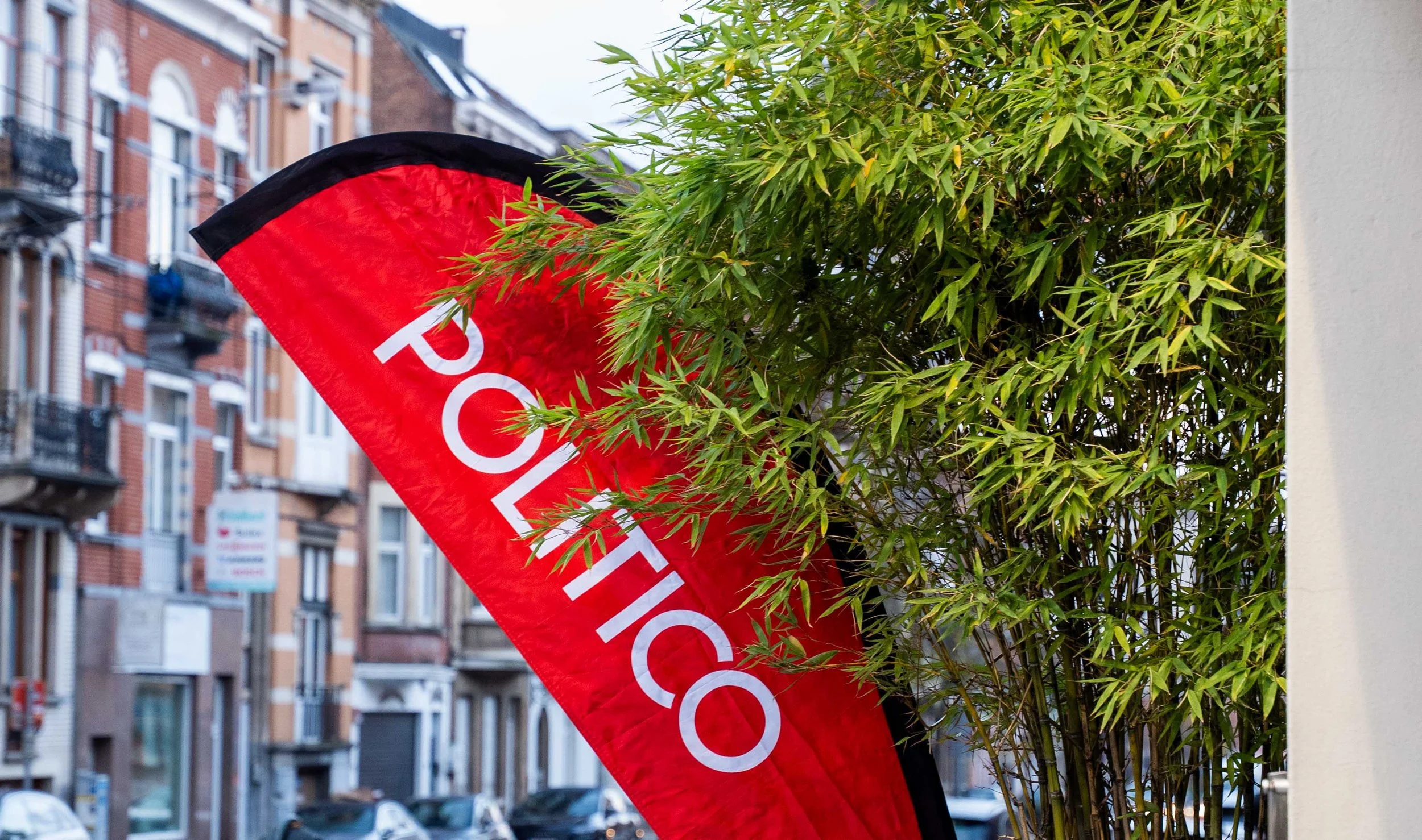 A red flag with white text that reads 'POLITICO', partially obscured by green bamboo leaves, on a city street with residential buildings in the background. Summit Photographer Brussels.