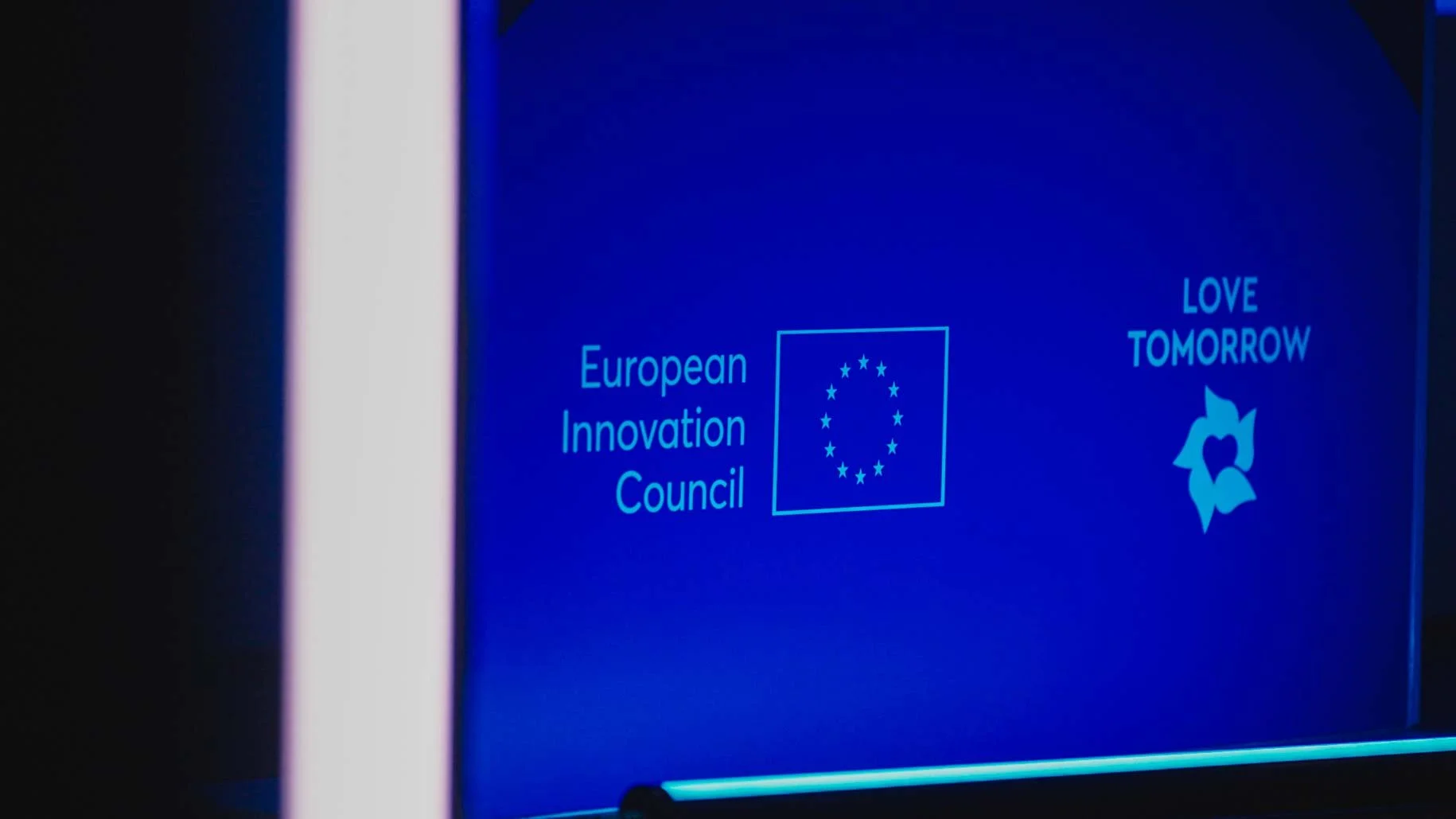 Blue screen with text reading 'European Innovation Council' next to the European Union flag, and a separate logo with the words 'LOVE TOMORROW' accompanied by a stylized flower design. Summit Photographer Antwerp & Brussels.