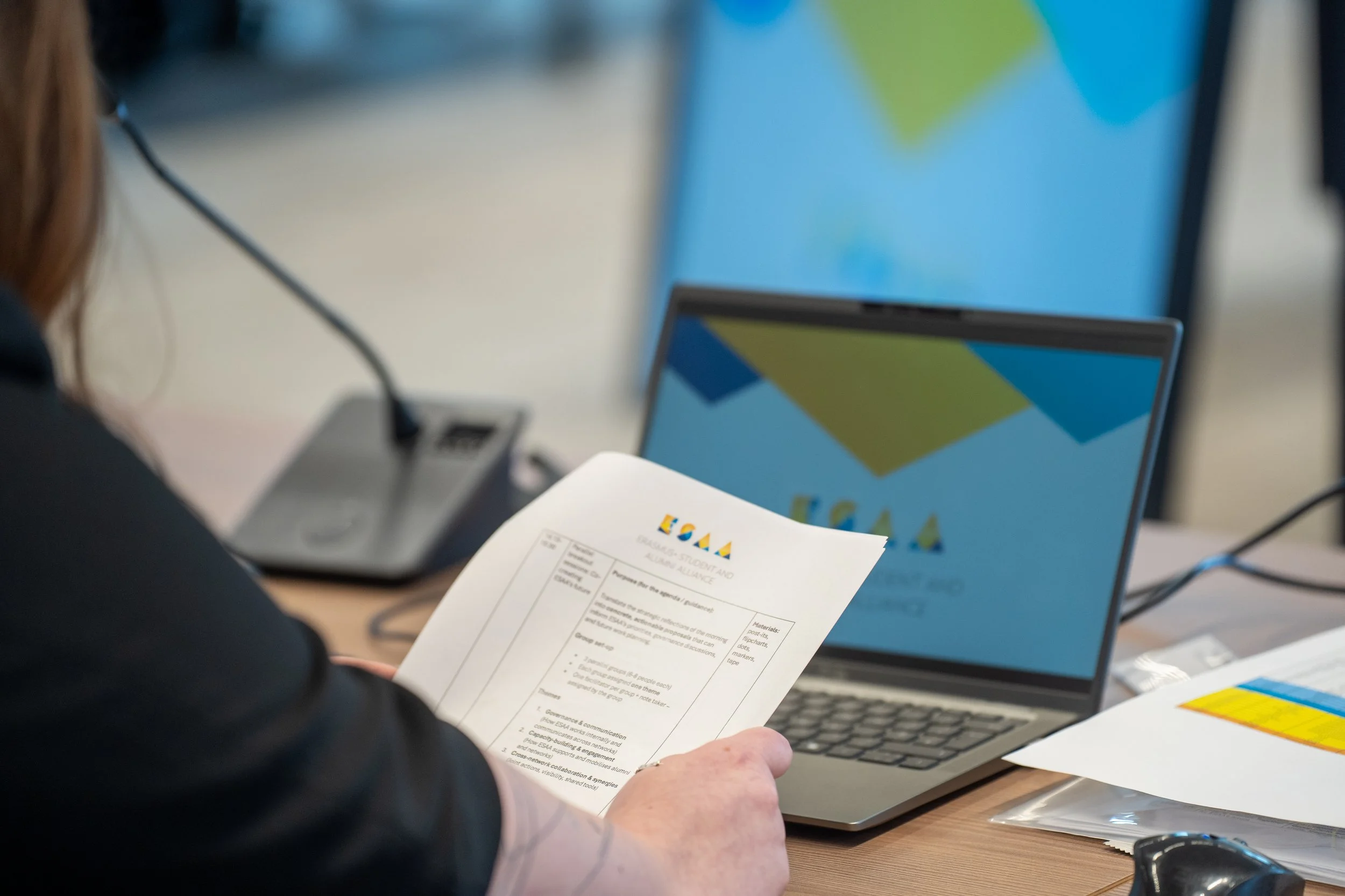 Person holding a printed document with a paper logo, seated at a desk with a laptop displaying the WordPress logo, connected via a black cable. Students of the Erasmus+ ESSA European Comission meeting in Brussels.